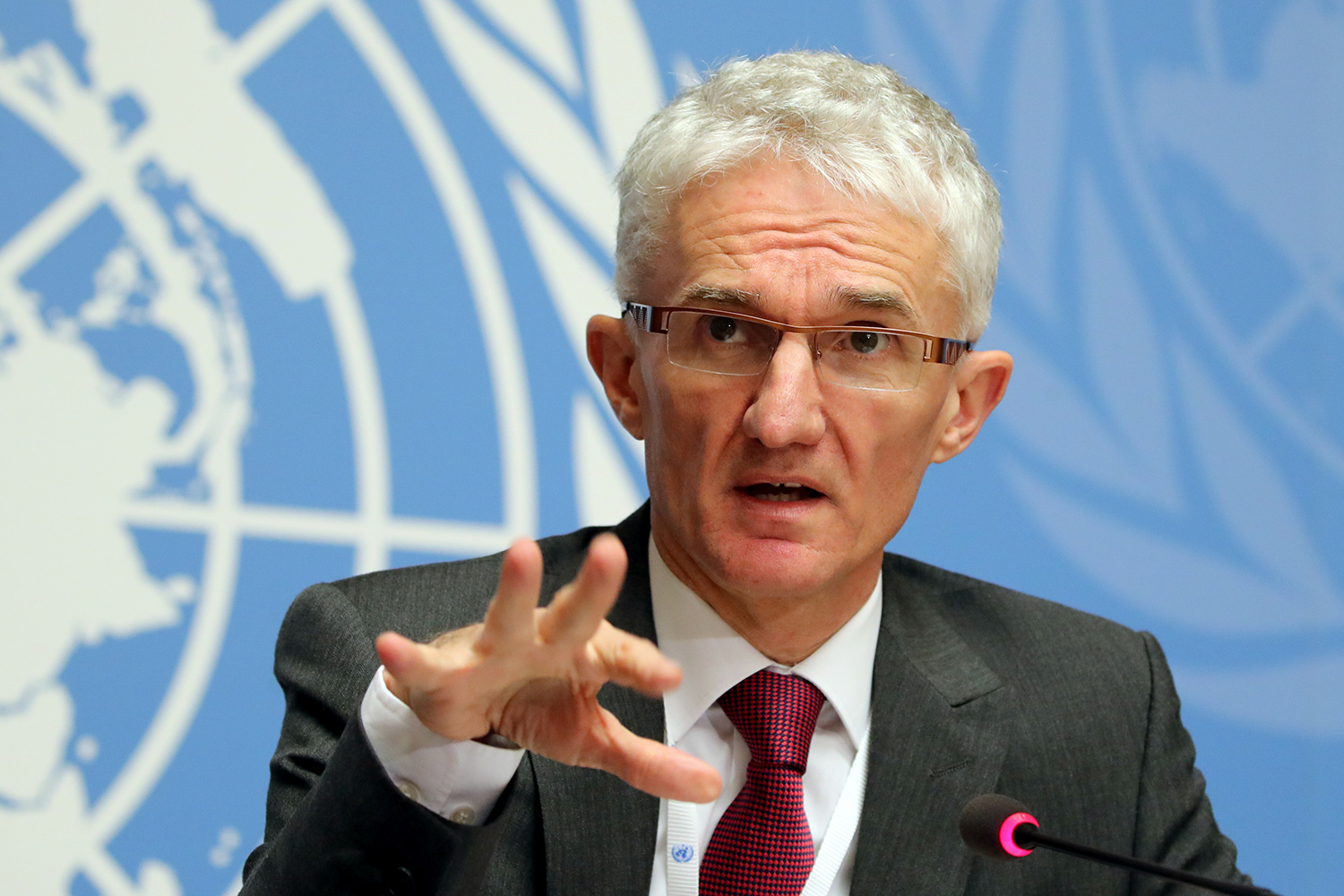 The official is seated at a table speaking into a microphone and gesturing with his hand. A blue and white UN logo is behind him.