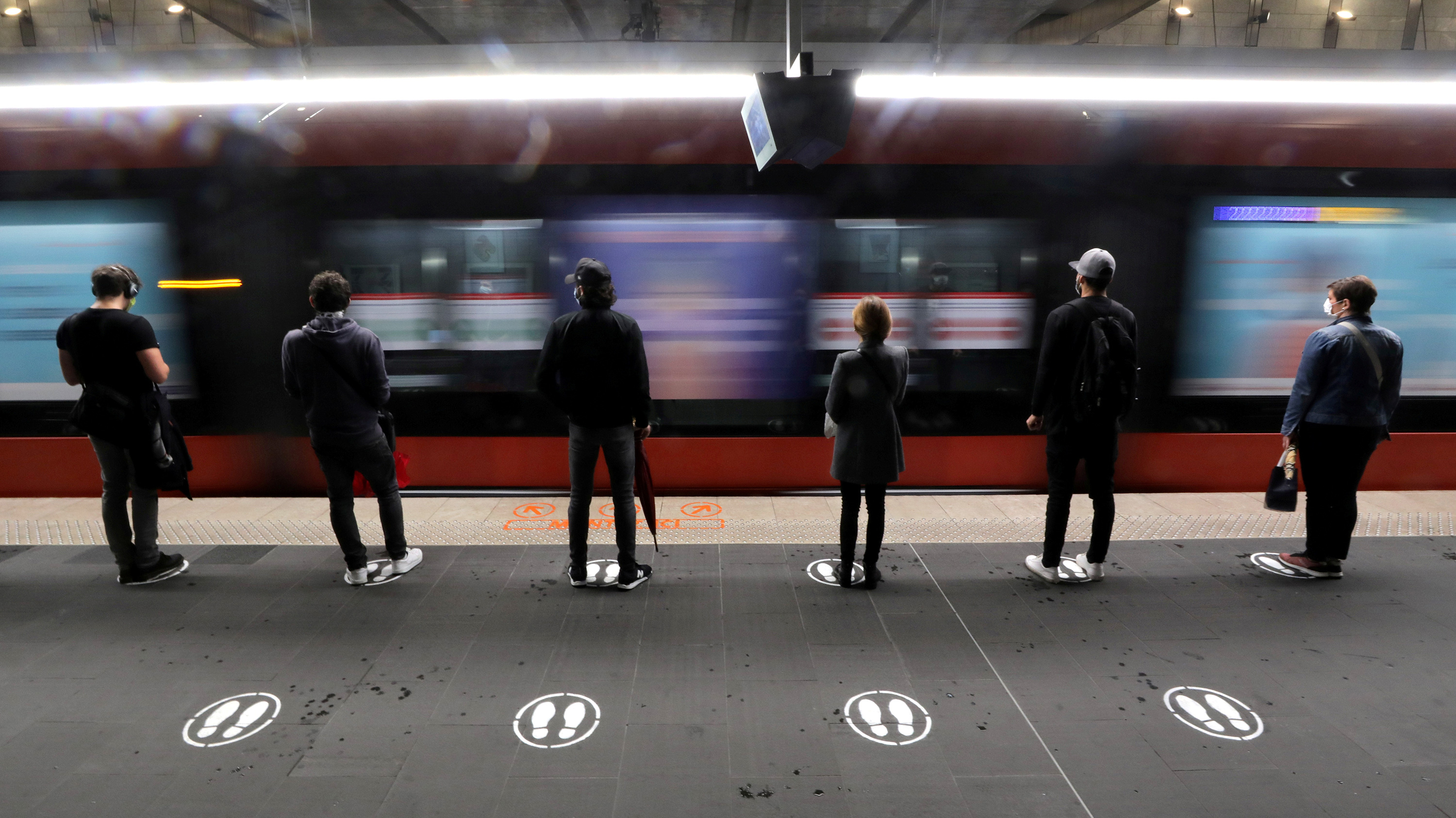 The photo shows people standing on paintef signs of feet on the platform with their backs to the camera as a train rushes by.