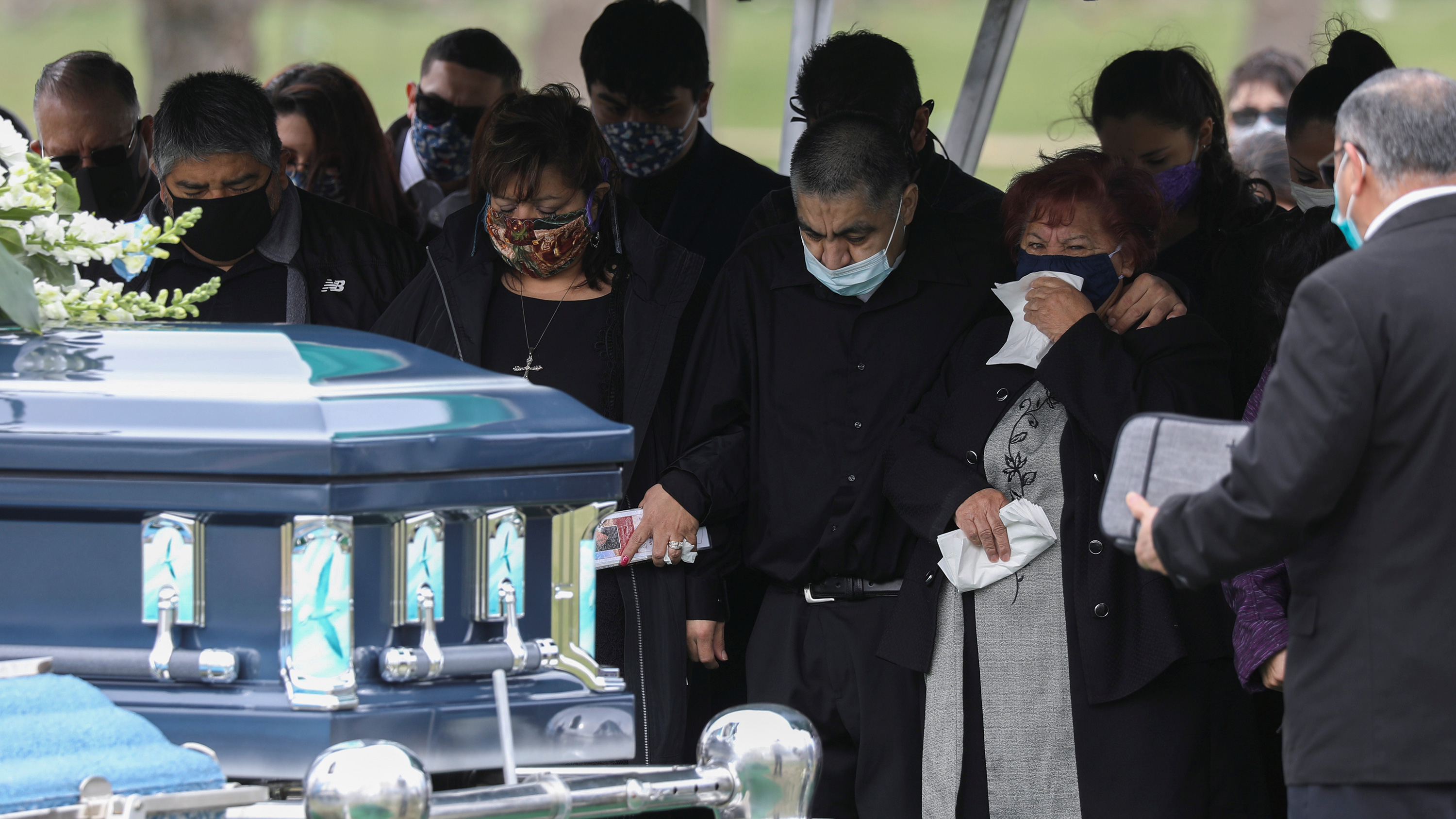 The photo shows a funeral with numerous people weeping for their lost loved one. A casket stands close to the camera, ready to be lowered into the ground.