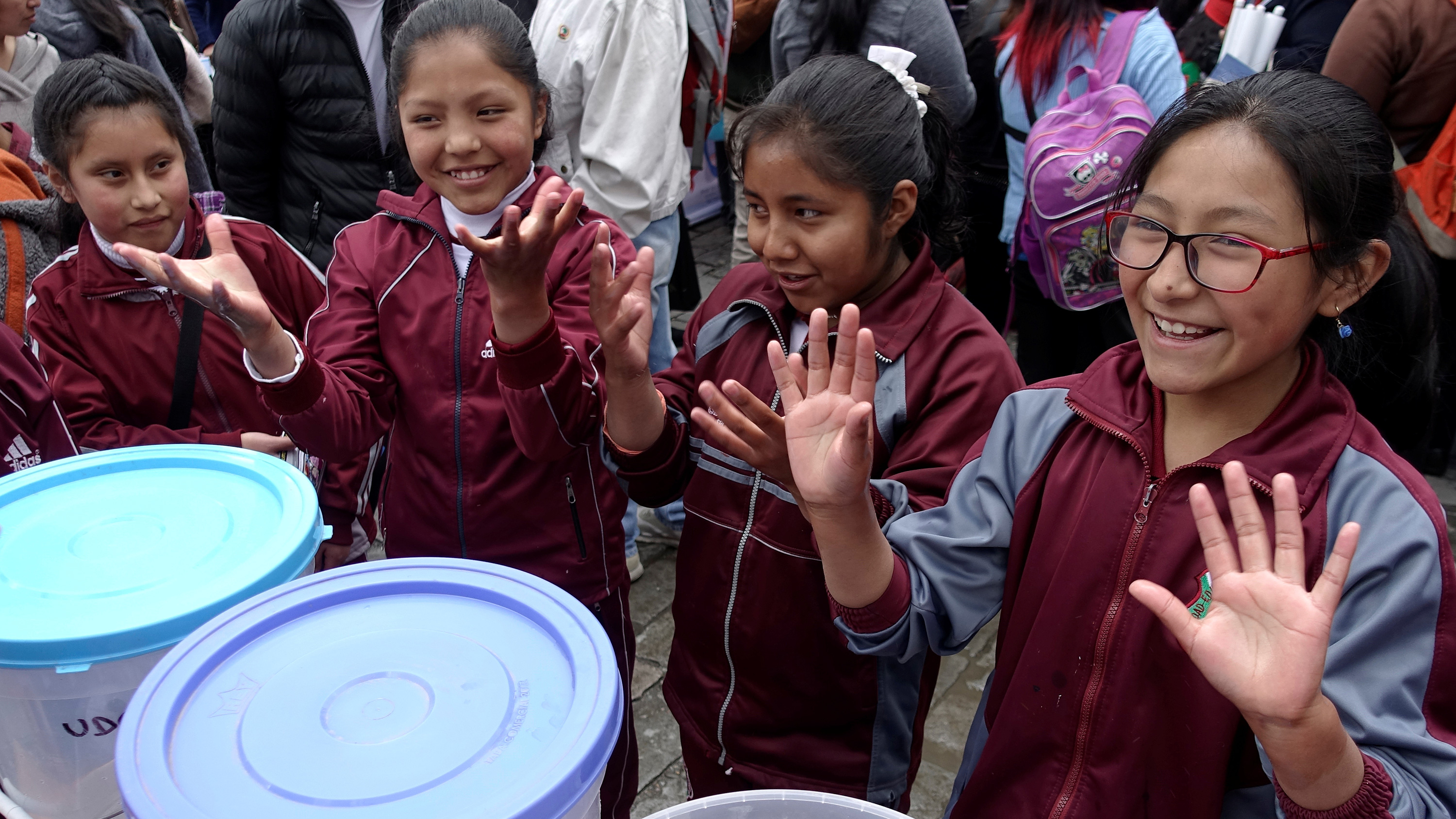 The photo shows a crowd of kids and adults. Four girls in school uniforms line up and show their hands to the camera.