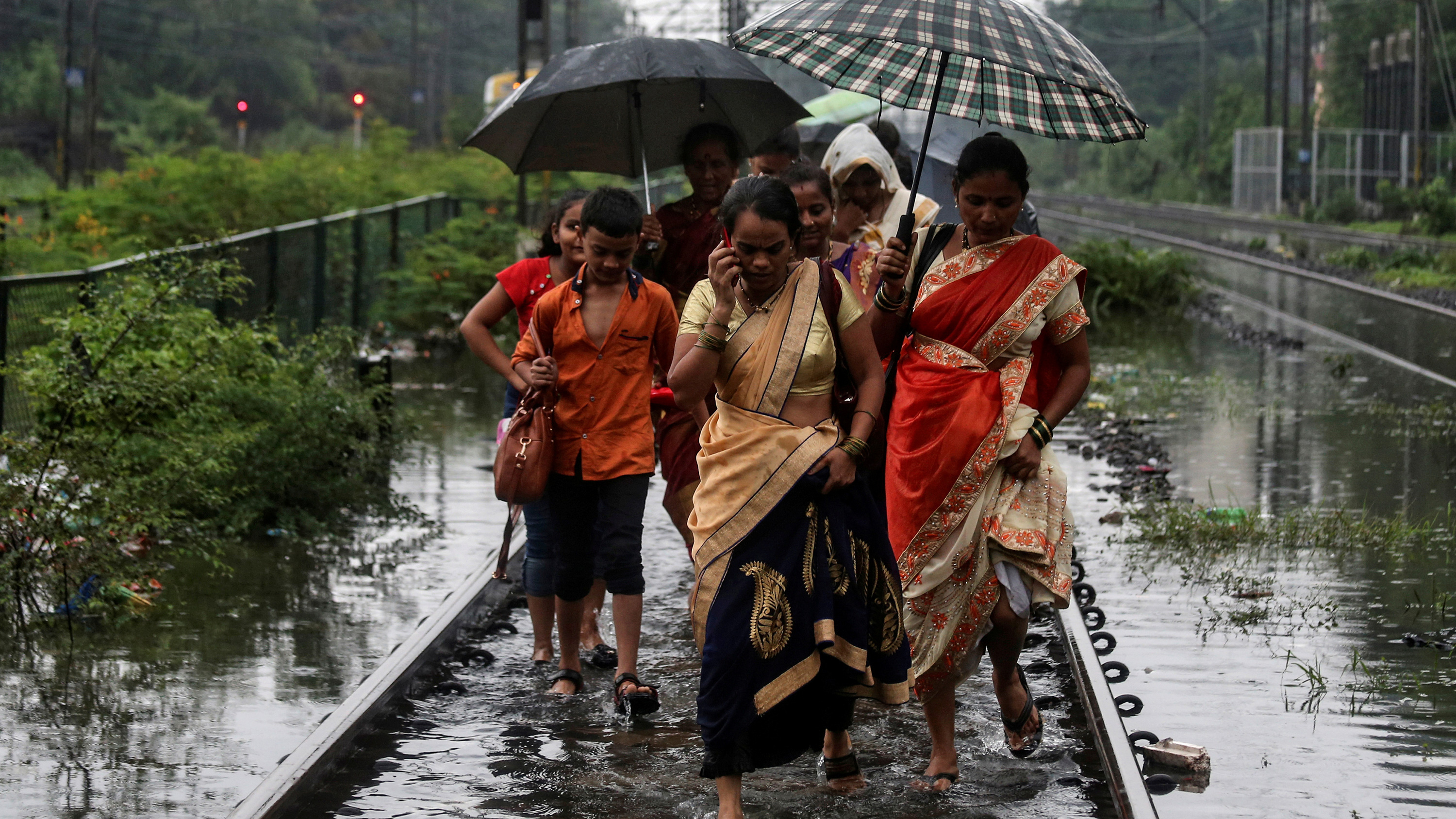 The image shows several people walking ankle-deep in water along railroad tracks as they hold several umbrellas over their heads in the rain.