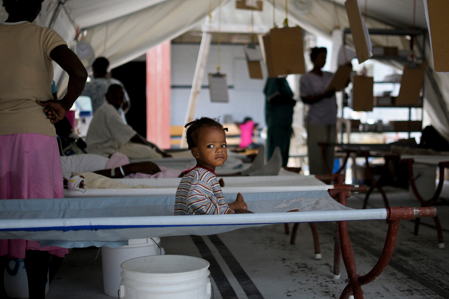 The photo shows a young child sitting up on a cot inside a tent with several other cots and people moving among them.