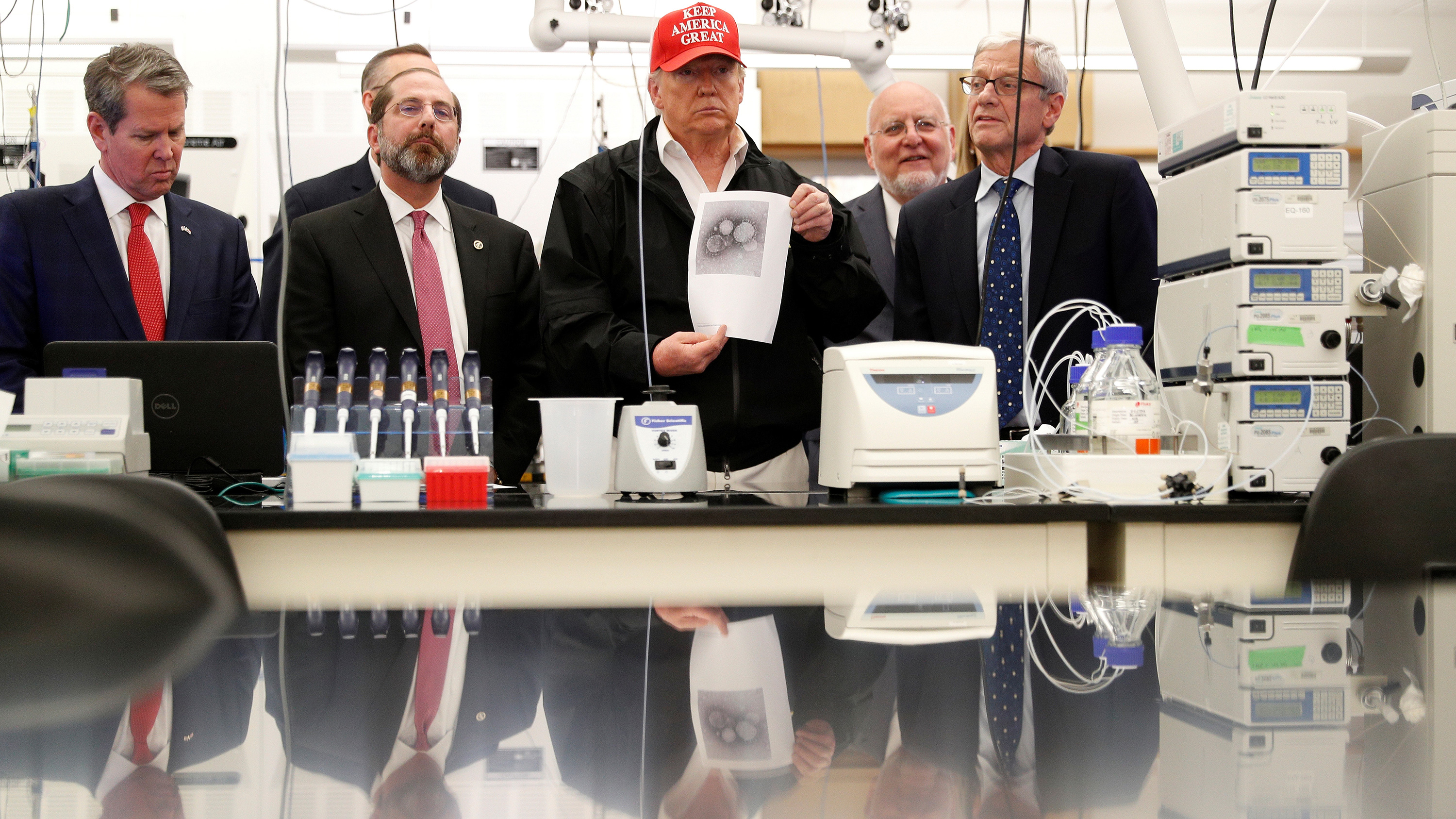 The photo shows President Trump and several others in a laboratory setting. The president is wearing a red "Make America Great Again" hat.