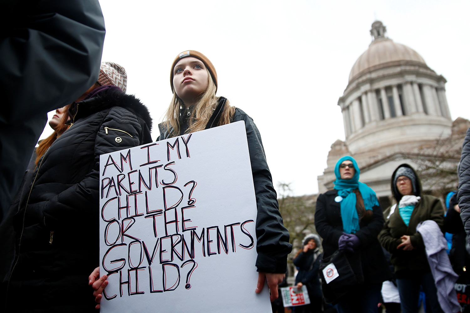The picture shows a girl holding a handmade sign that reads, "Am I my parent's child or the government's child?"