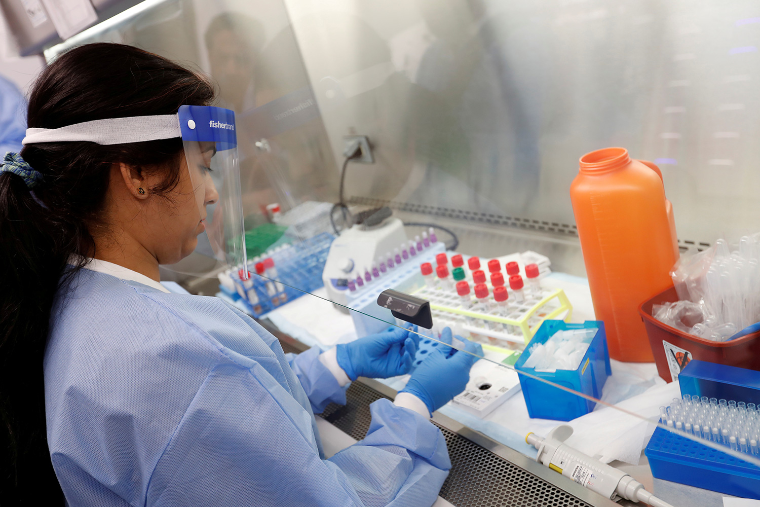 The photo shows a technologist working with vials and a pipette under a hood.