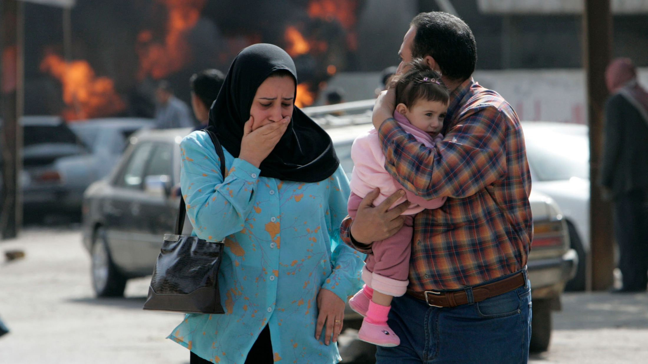 This is a powerful photo showing a woman choked up walking away from a fiery blaze of a car burning. Next to her is a man holding a small child. He is looking back on the scene of carnage.
