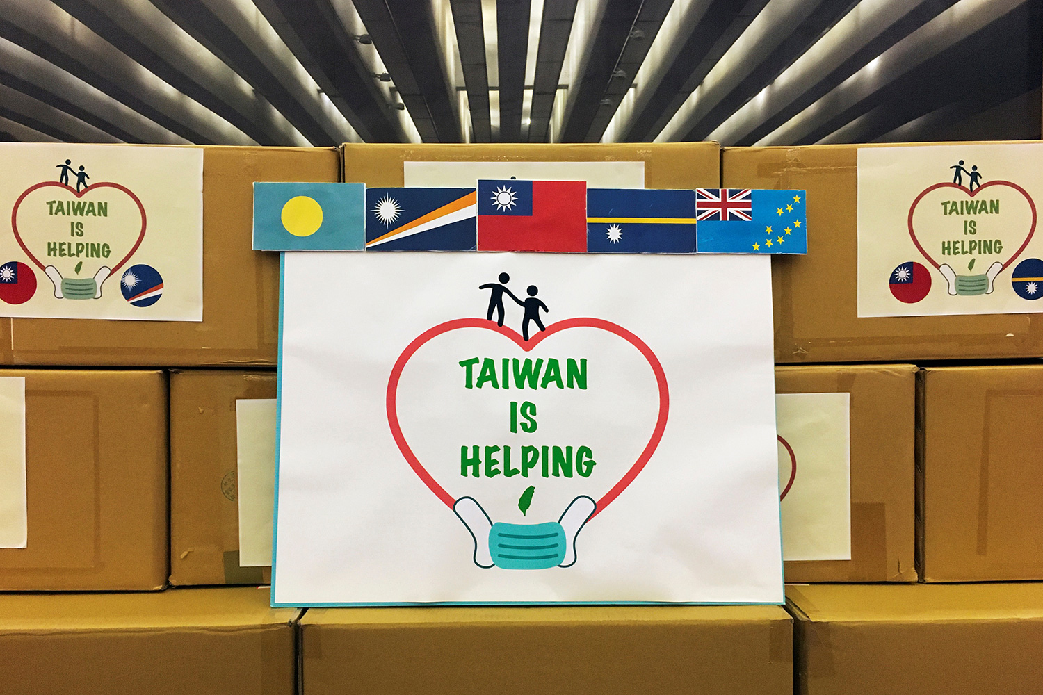 The photo shows boxes stacked and decorated with country flags and a large sign that reads, "Taiwan Is Helping."