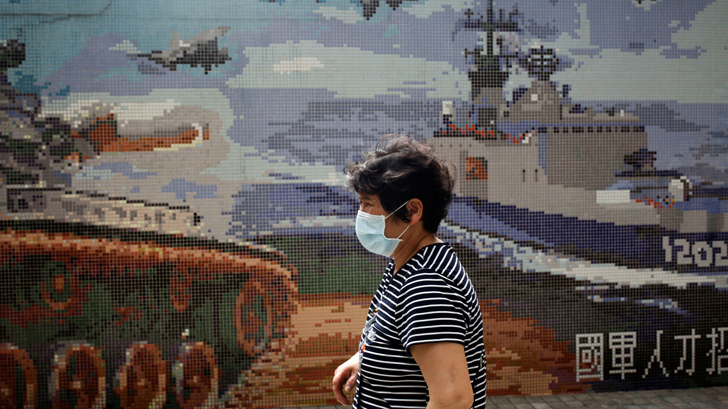 The photo shows a woman in a mask walking past a stylized poster showing war tanks, ships and planes.