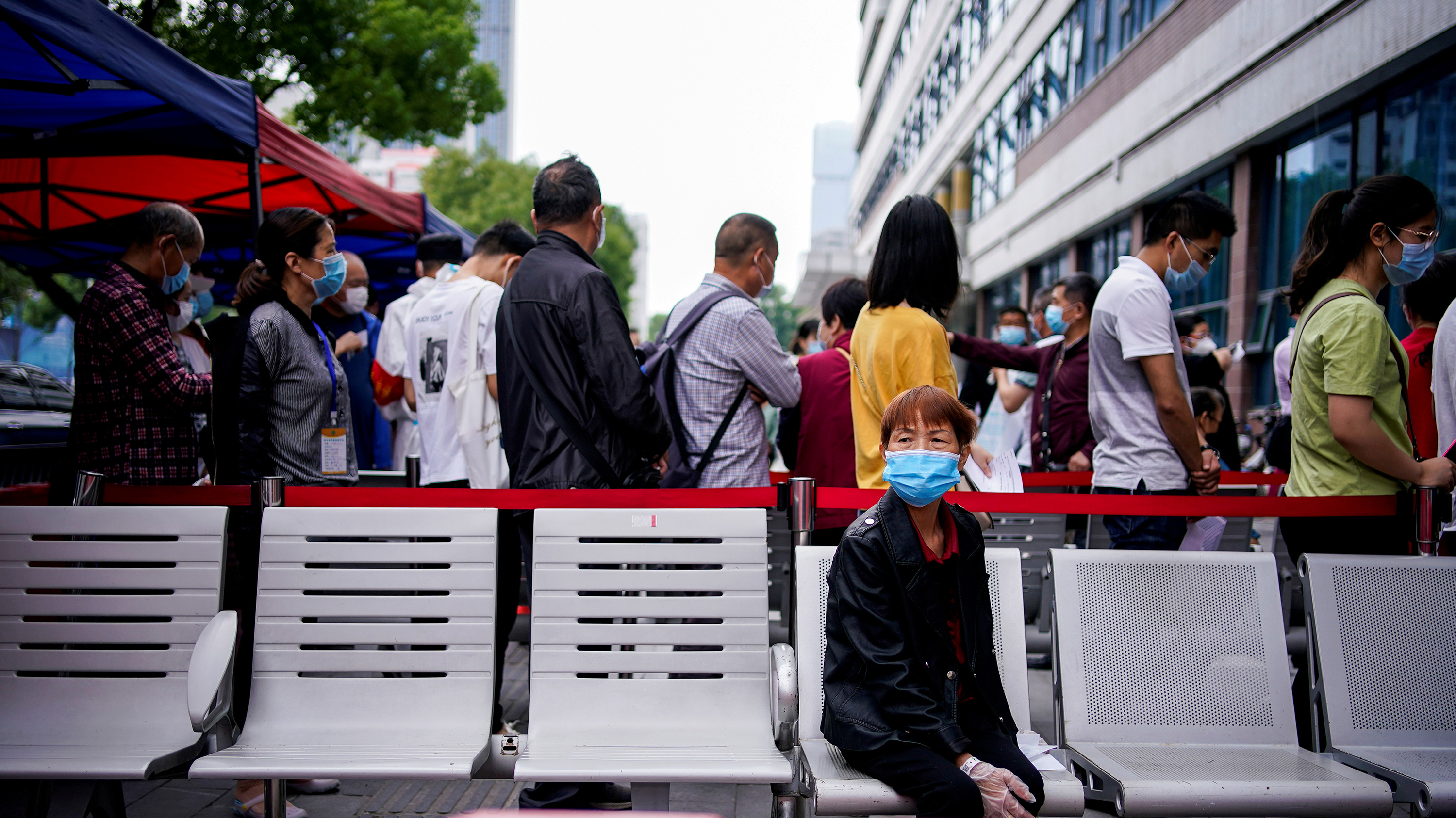 The photo shows a woman sitting on the back side of a barricade along which people are lined up.
