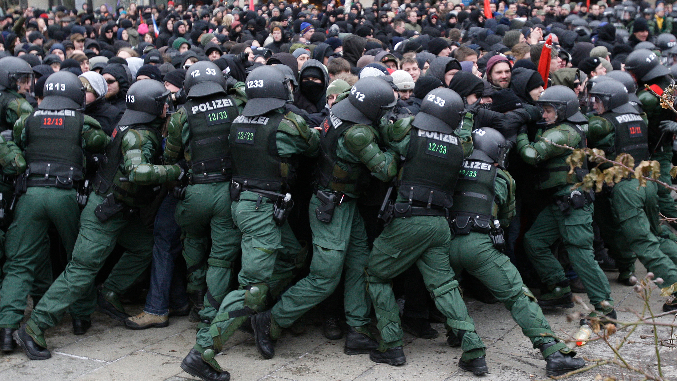 The photo shows a line of uniformed riot gear officers pushing hard against a scrum of protesters.