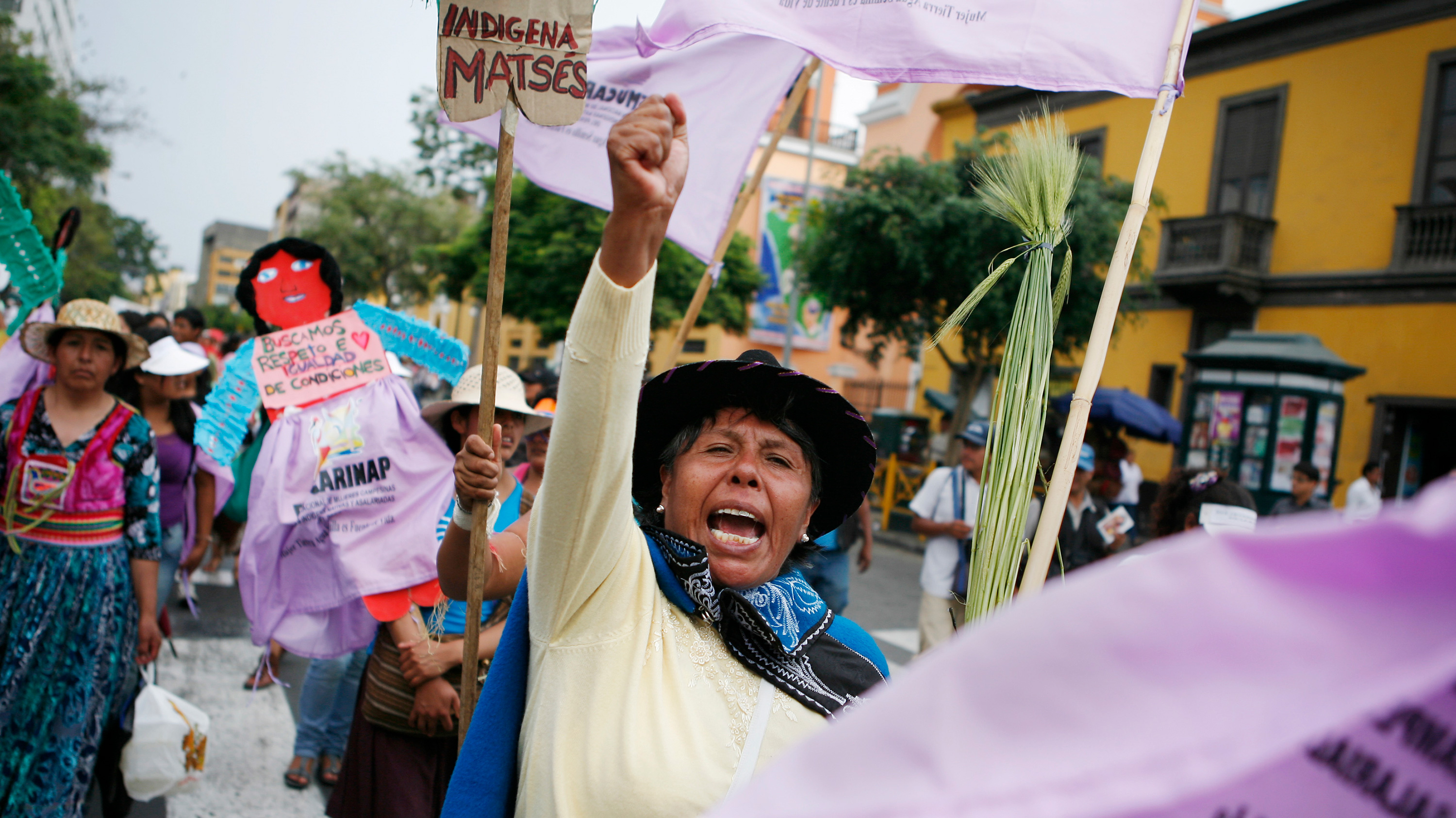 The photo shows a line of protesters marching down a street with a woman in the middle of the frame raising her hist and shouting.