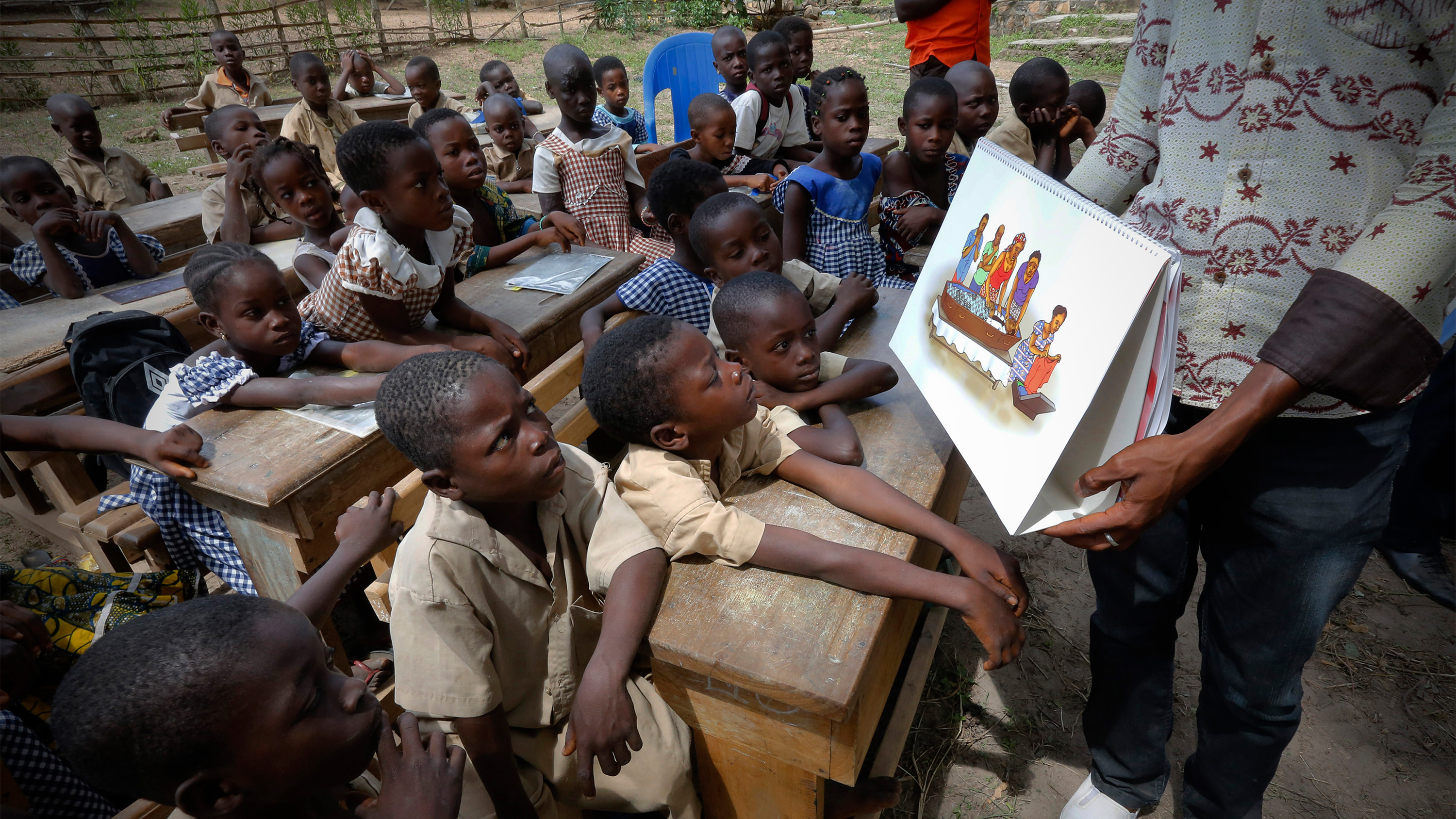 The photo shows the teacher at the front of a room showing a placard to the children.