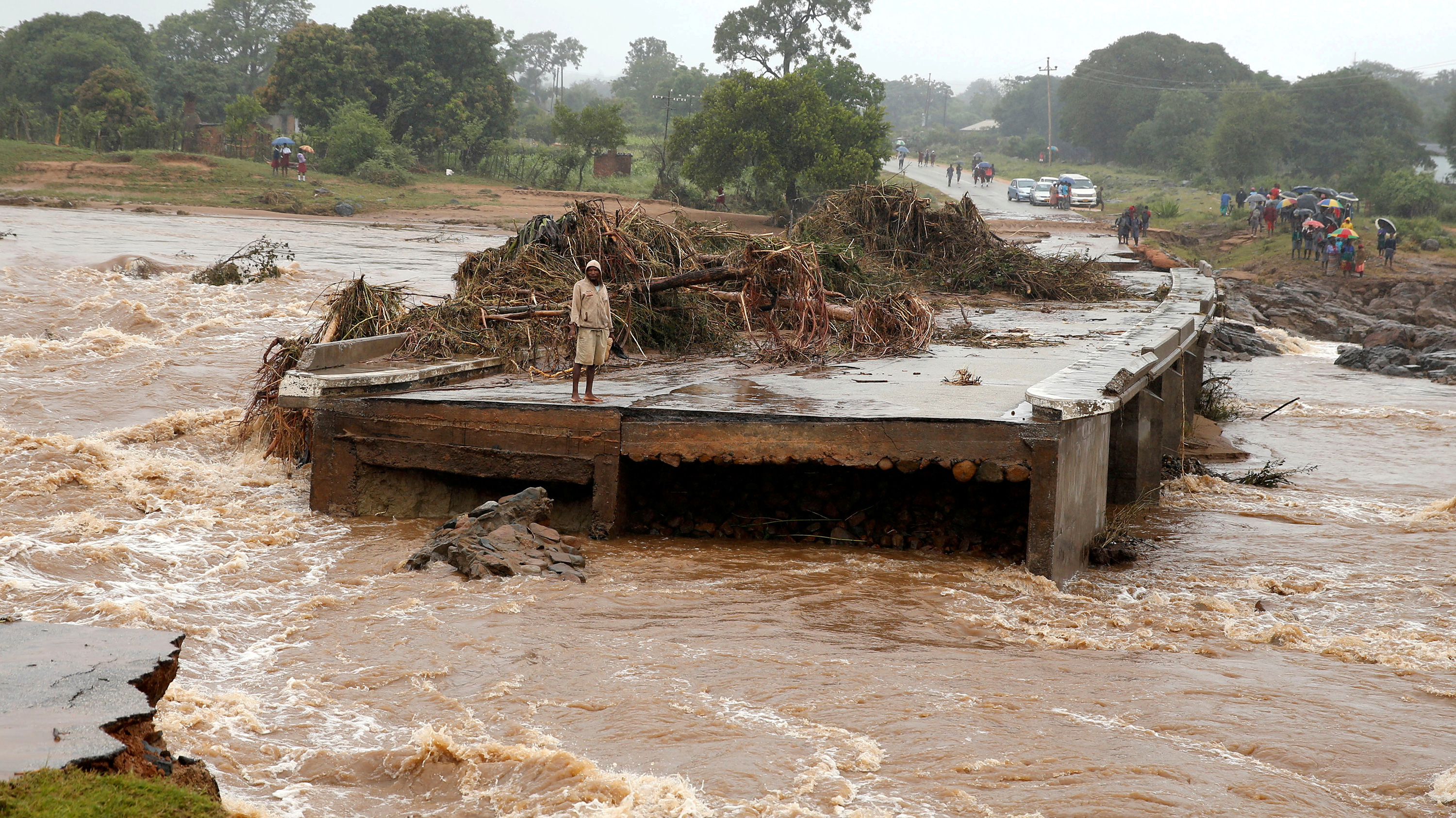 The photo shows a man at one end of a bridge that is completely washed away from the flooding.