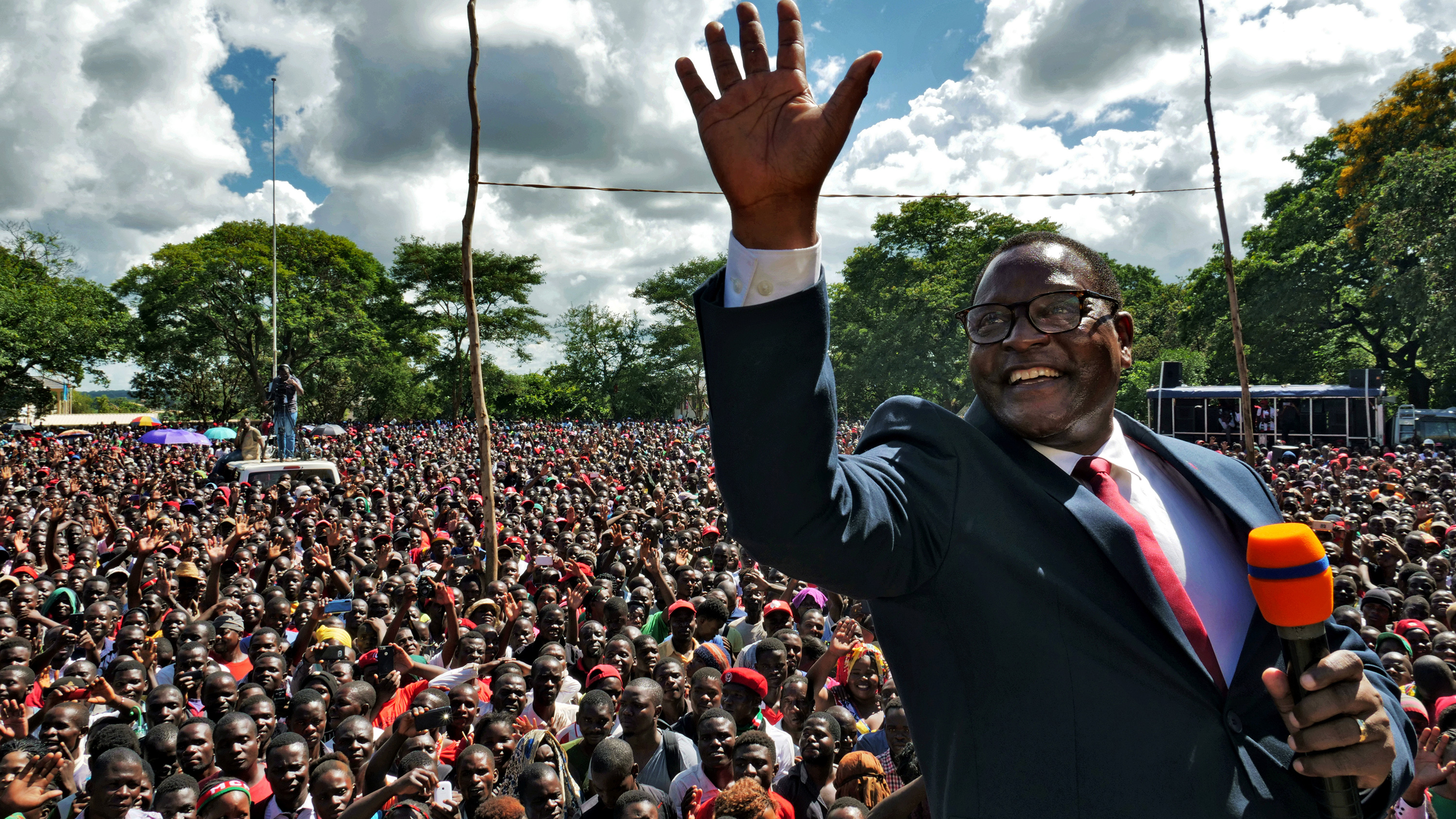 The photo shows a smiling Chakwera, microphone in hand, standing atop a stage waving and smiling. A massive crowd can be seen in the background.