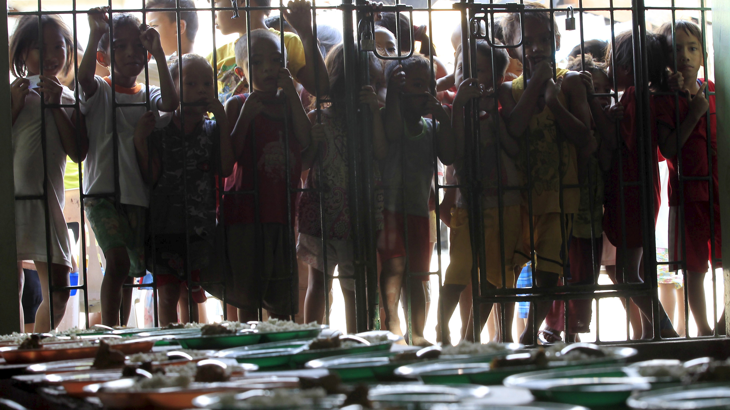 The photo shows children lined up along the bars keeping them from the interior of the space wherein the photographer is standing. In the foreground can be seen a table piled with individual meals.