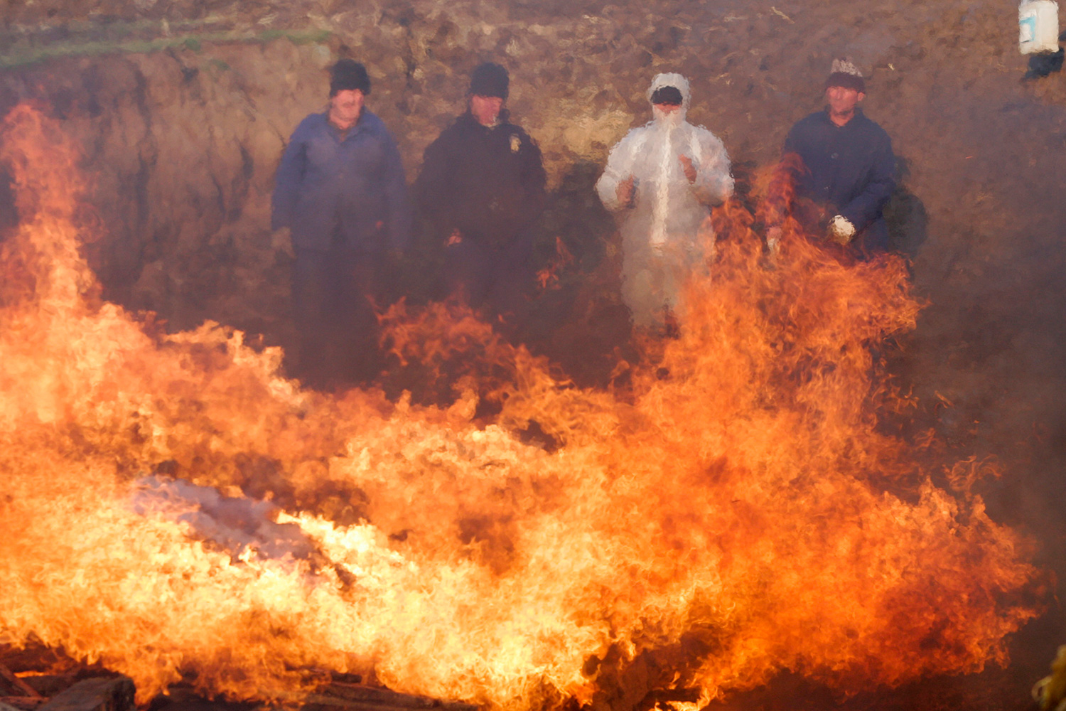 The photo shows three people wearing protective suits standing behind a huge fire.