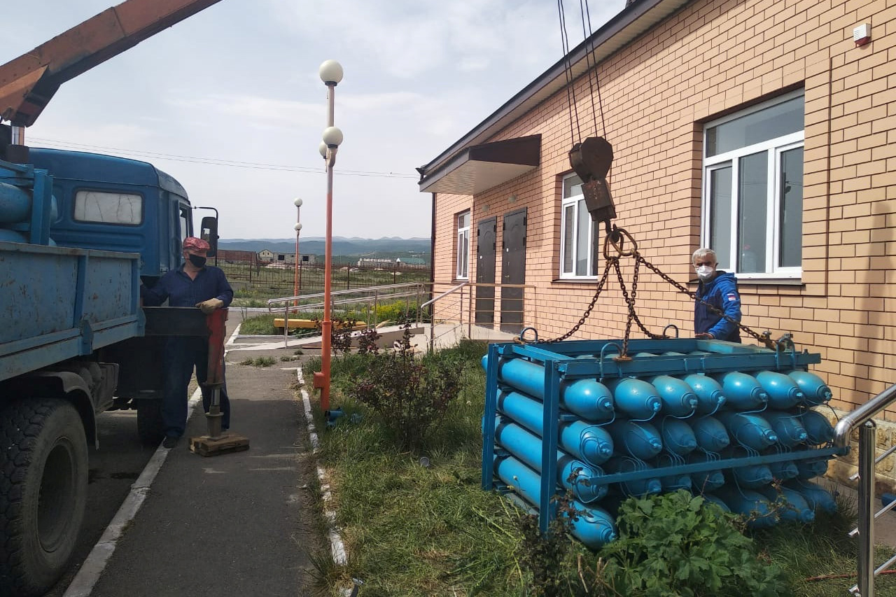 Photo shows two workers with a truck that has a crane arm attached to a large palette of bright blue, 5-ft long oxygen tanks.