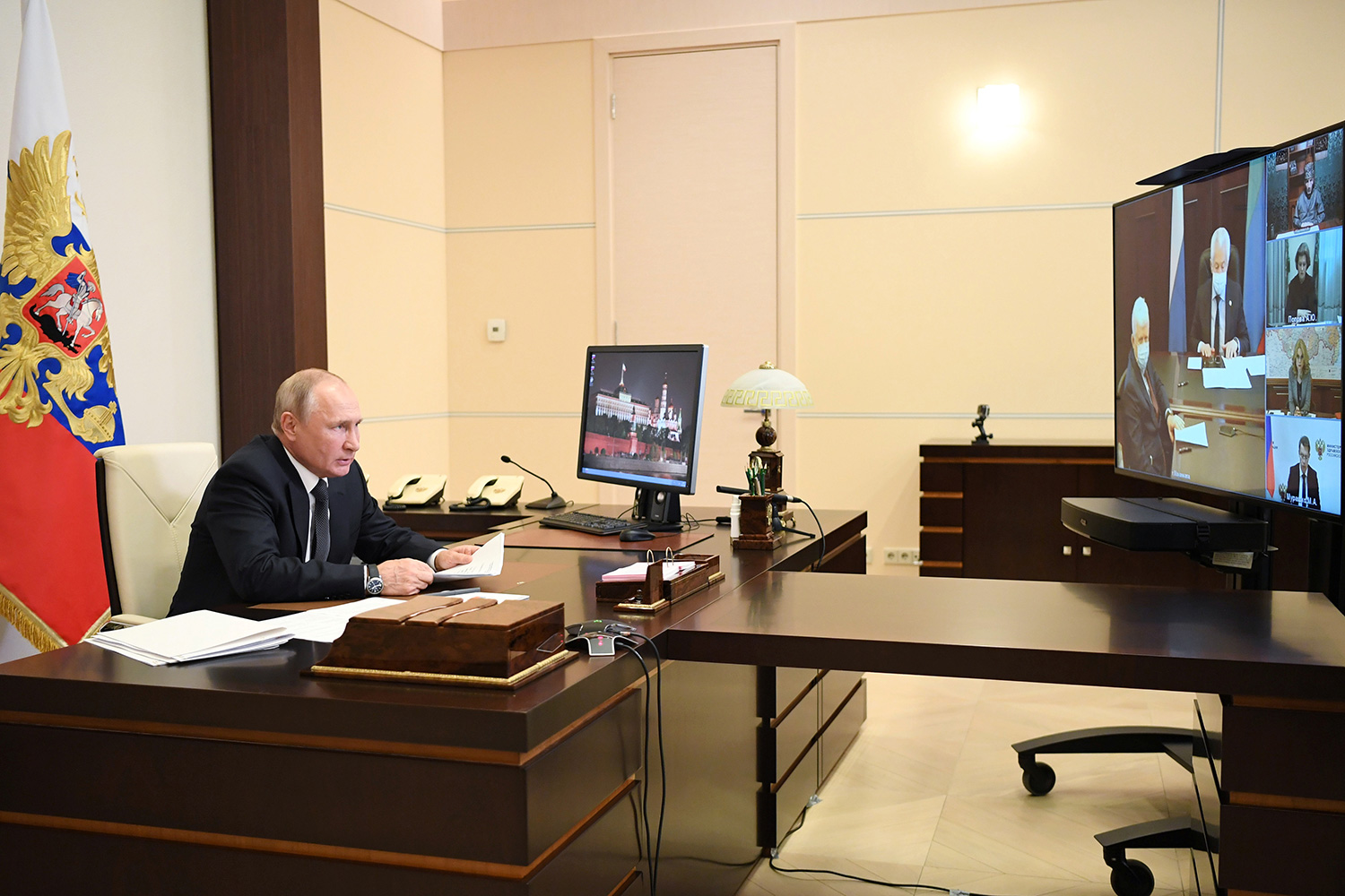 The image shows Putin at a desk across from a large screen on which others can be seen conferenced in.