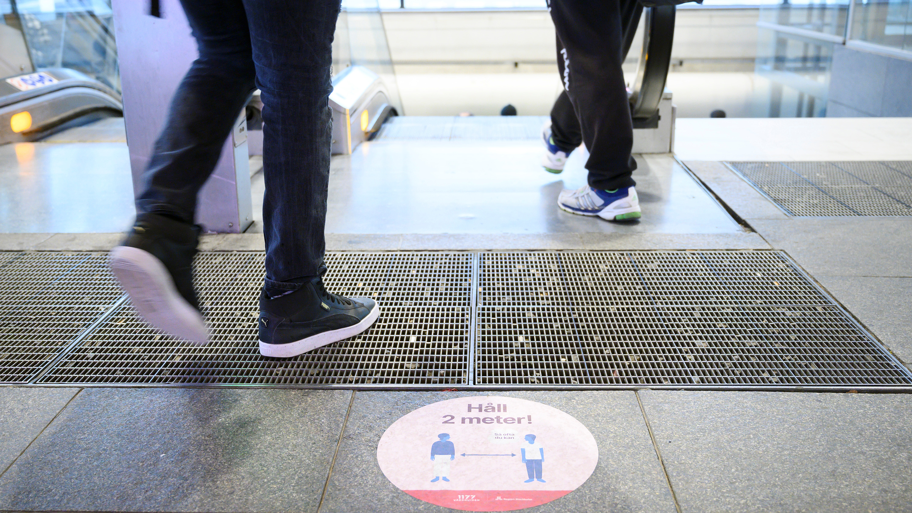The photo shows feet walking onto an escalator above a sign warning of social distancing.