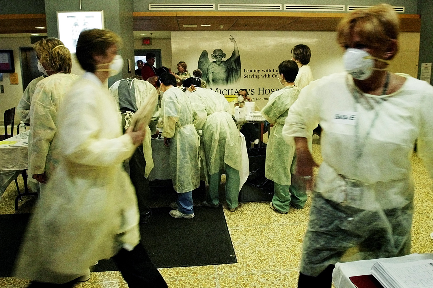 The photo shows a bustle of hospital staff moving around the waiting area of a hospital, whose namesake is the subject of a mural in the background, behind the information desk.