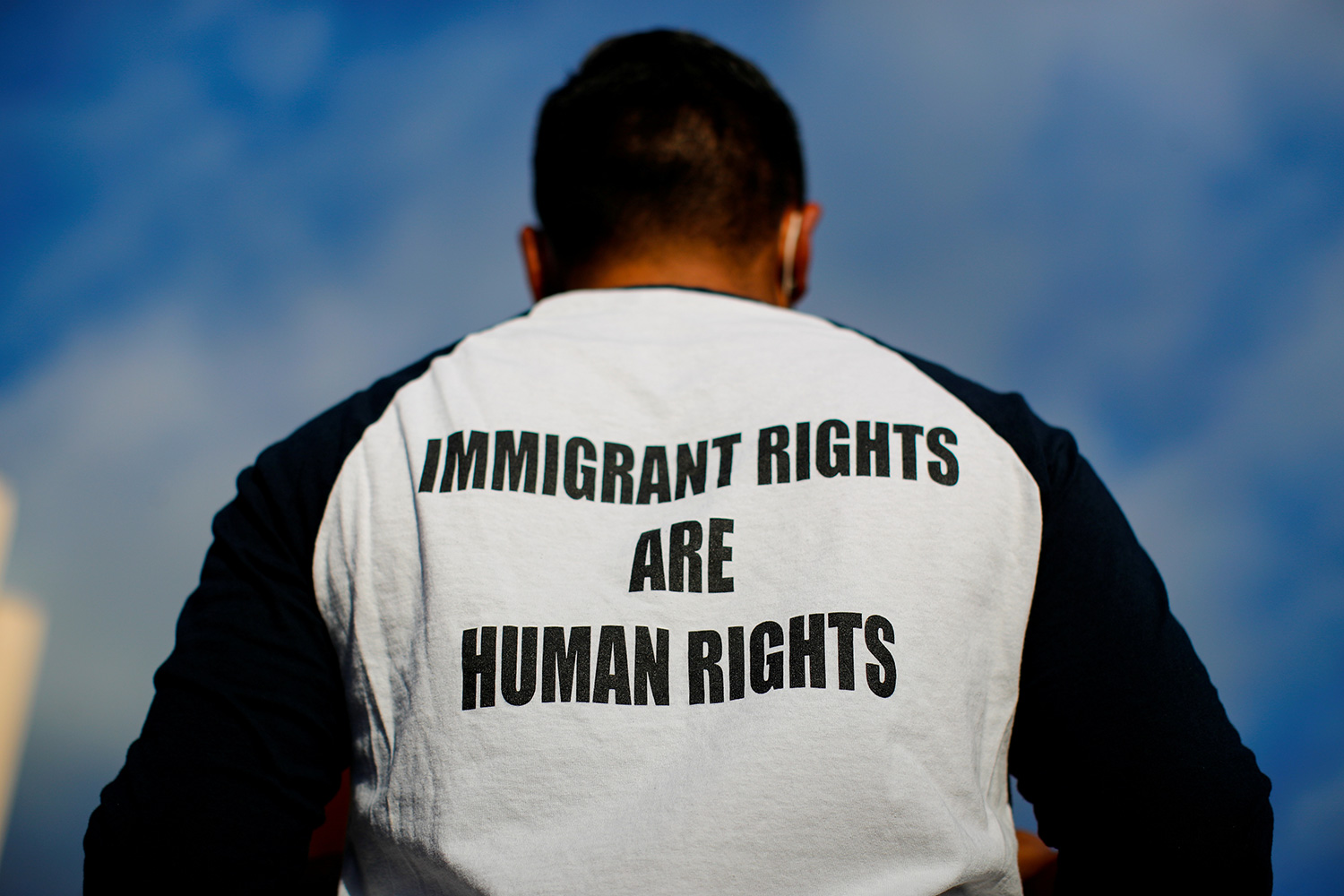 The photo shows a man with his back to the camera wearing a black-and-white baseball shirt emblazoned with the message, "Immigrant rights are human rights."