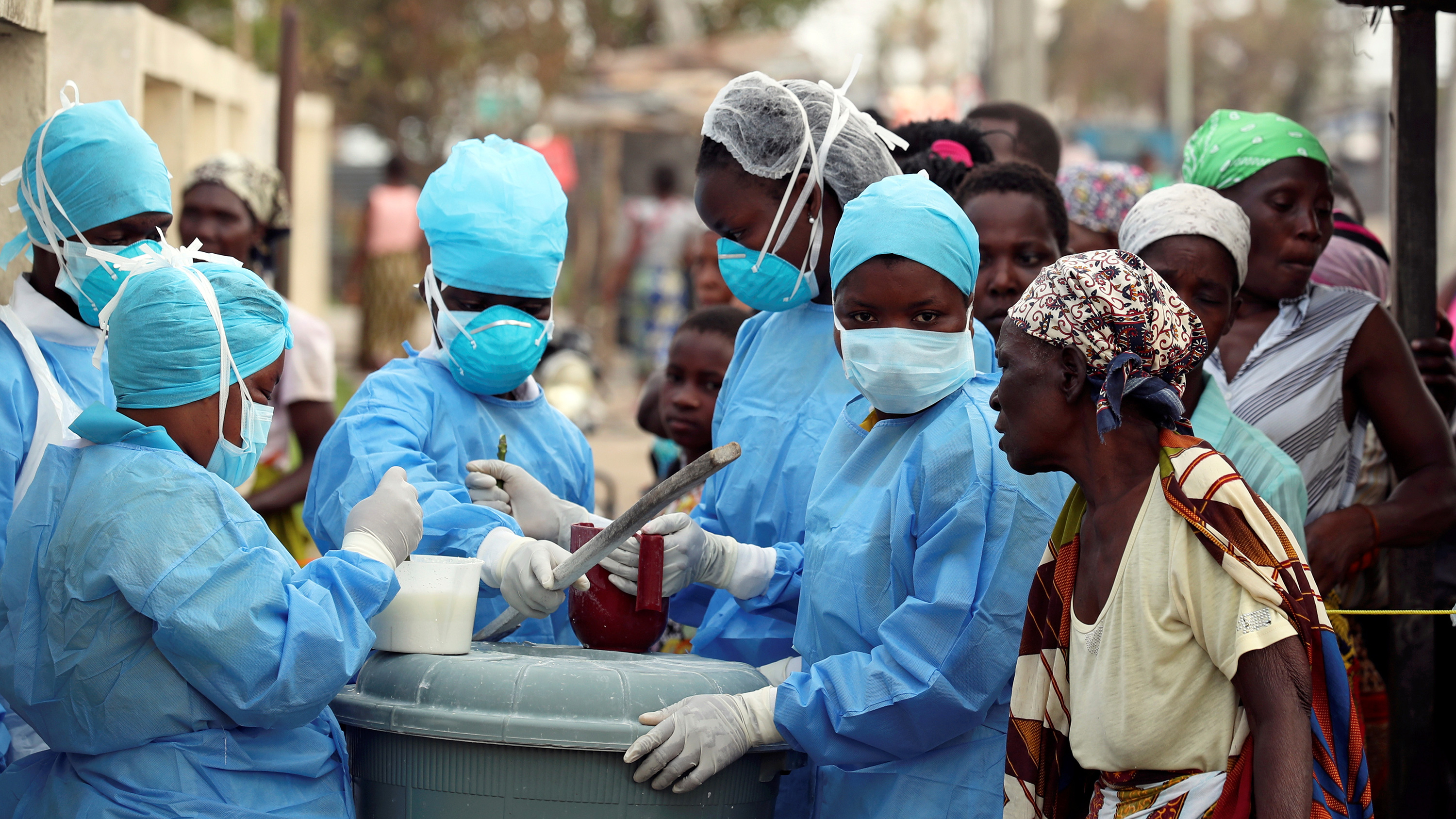 The photo shows a number of people wearing blue protective gear working on mixing a solution with a number of others wearing plain clothes looking on.