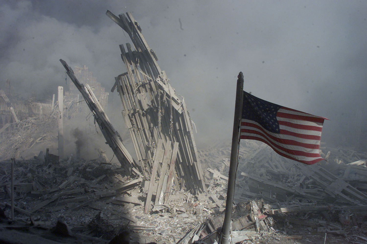 The photo shows a piece of the tower base shorn off at ground level with the American flag waving through the grim haze in the foreground.