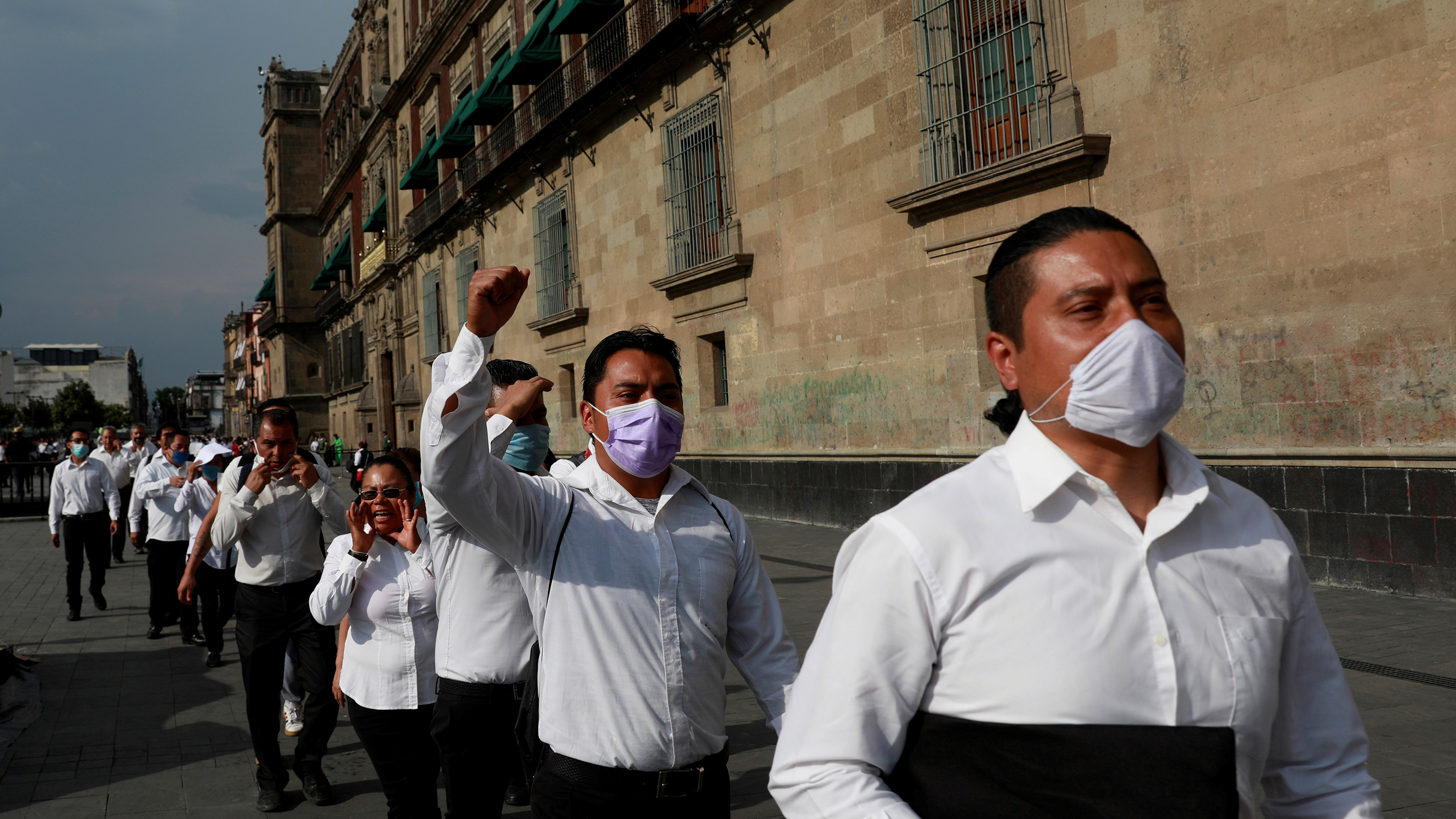 Photo shows a line of marching protestors wearing wait staff whites and angry looks. Fists raised, several are wearing masks.