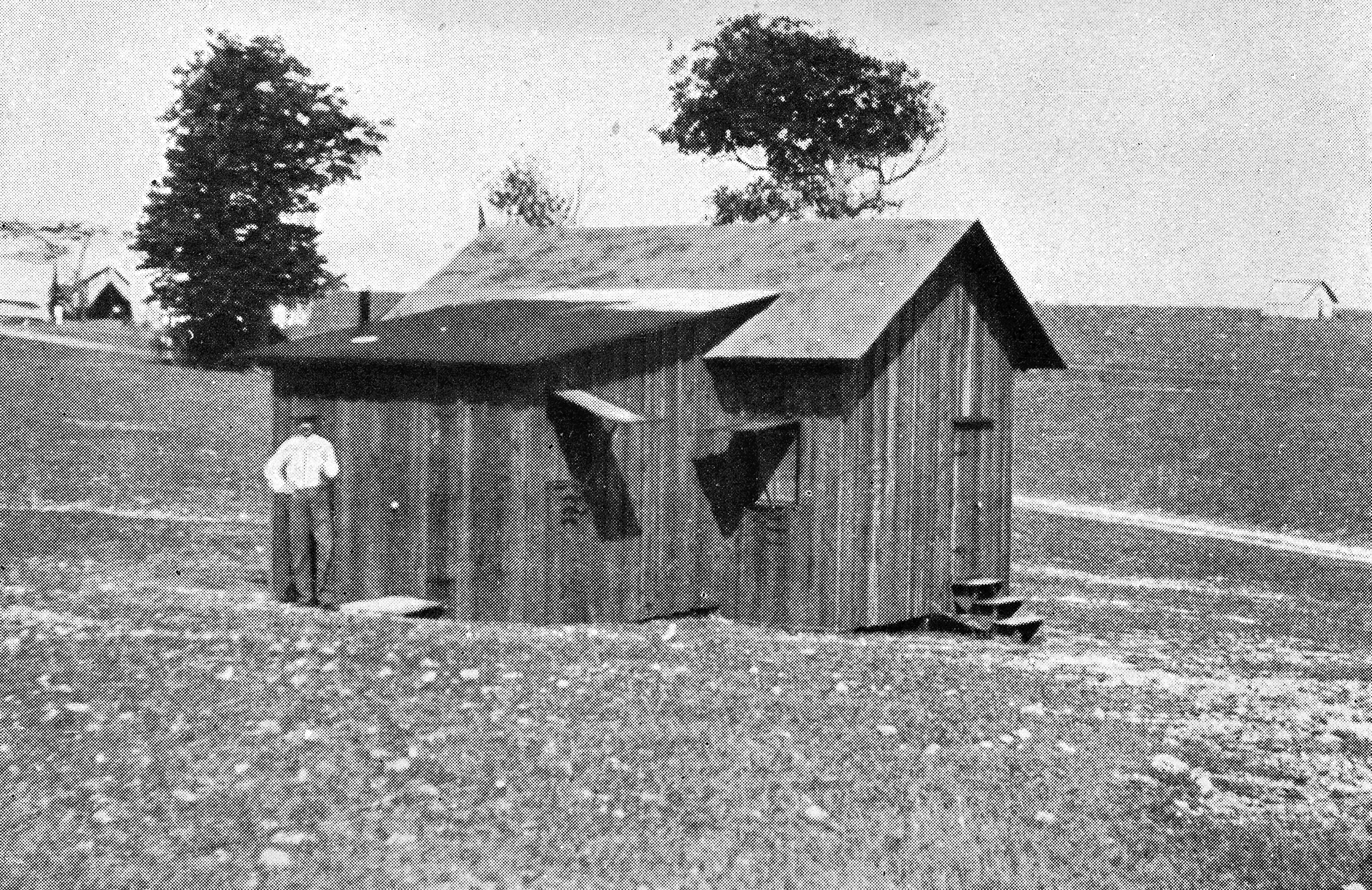 The historic photo shows a wooden hut of a building in a desolate setting with a man standing out front.