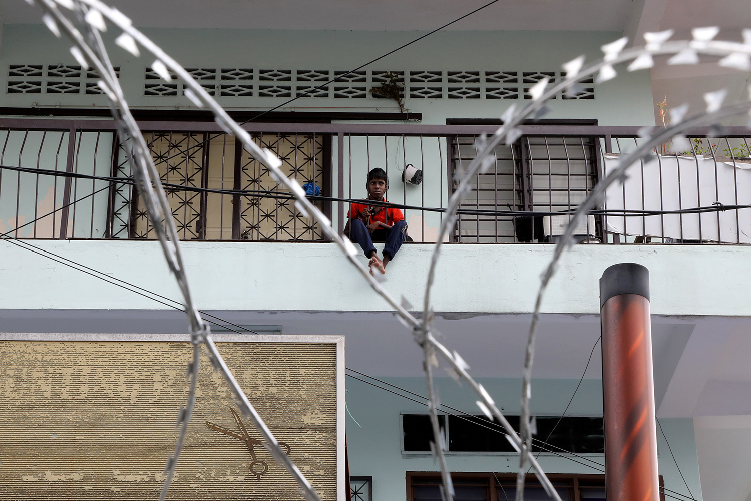 The photo shows the boy from a distance, standing on a balcony with barbed wire seen in the foreground.