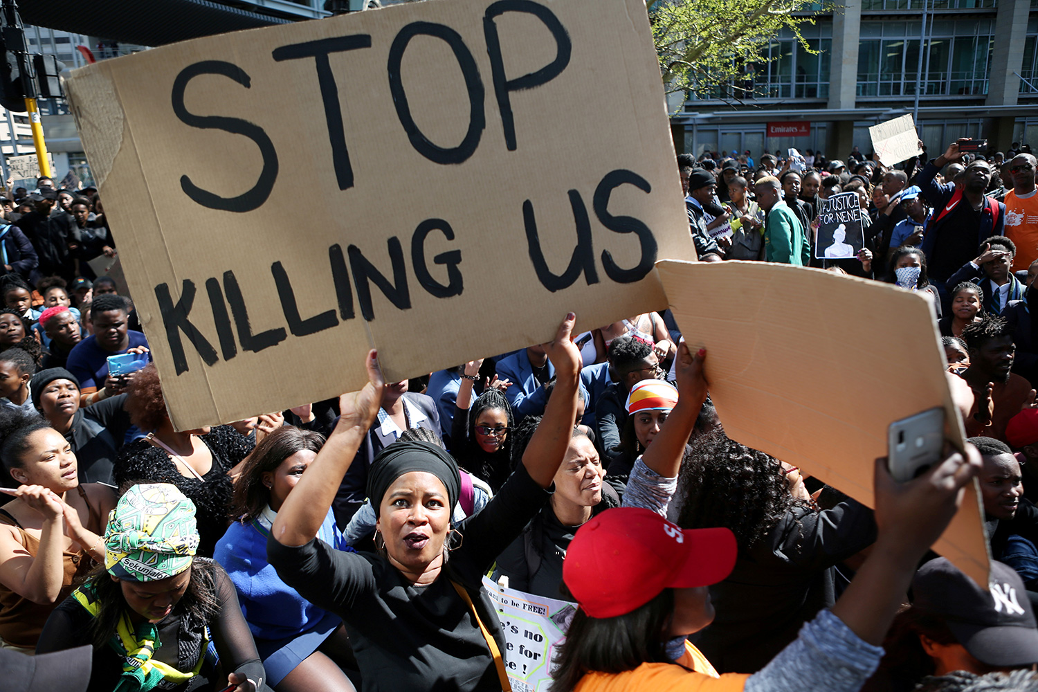 The photo shows a large crowd of protestors with one woman in the foreground holding up a large sign that reads, "Stop Killing Us."