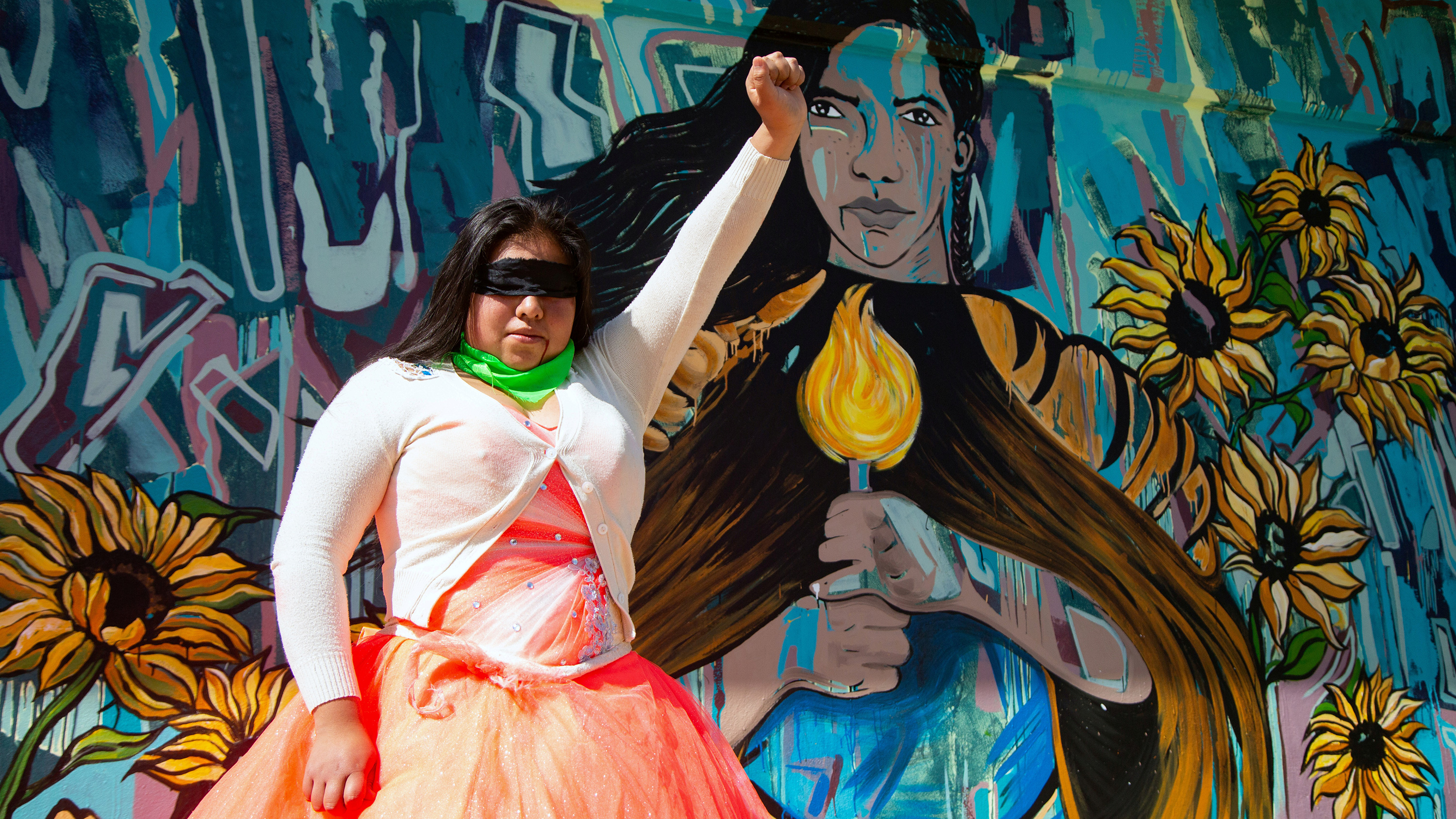 The photo shows a woman in an orange dress and white cardigan with a blindfold over her face holding her fist in the air while she stands in front of a painted mural of a woman holding a candle.