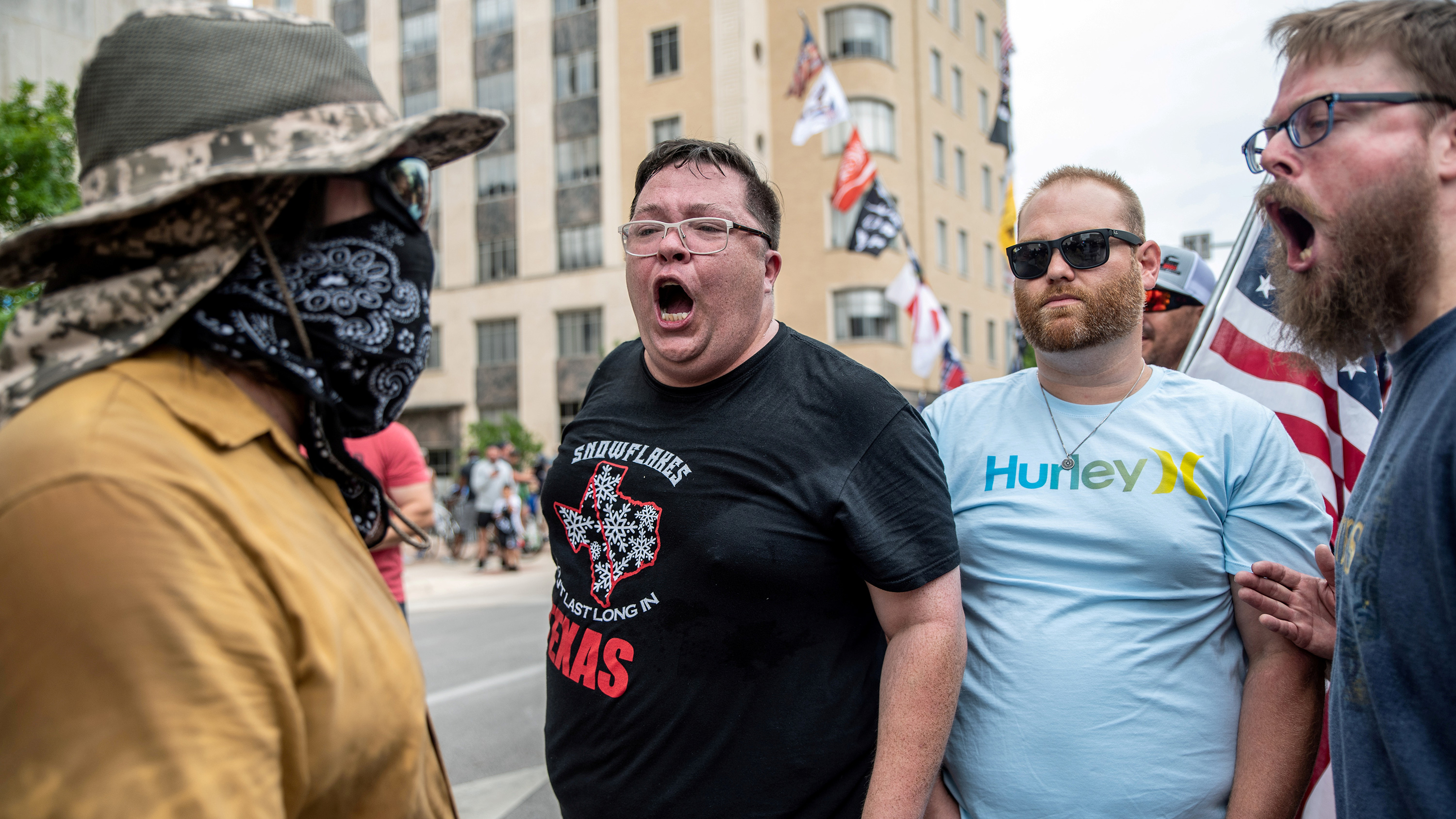The picture shpws a standoff between three non-mask wearing protesters shouting at a single protest counterpart who has a bandana fashioned as a mask.