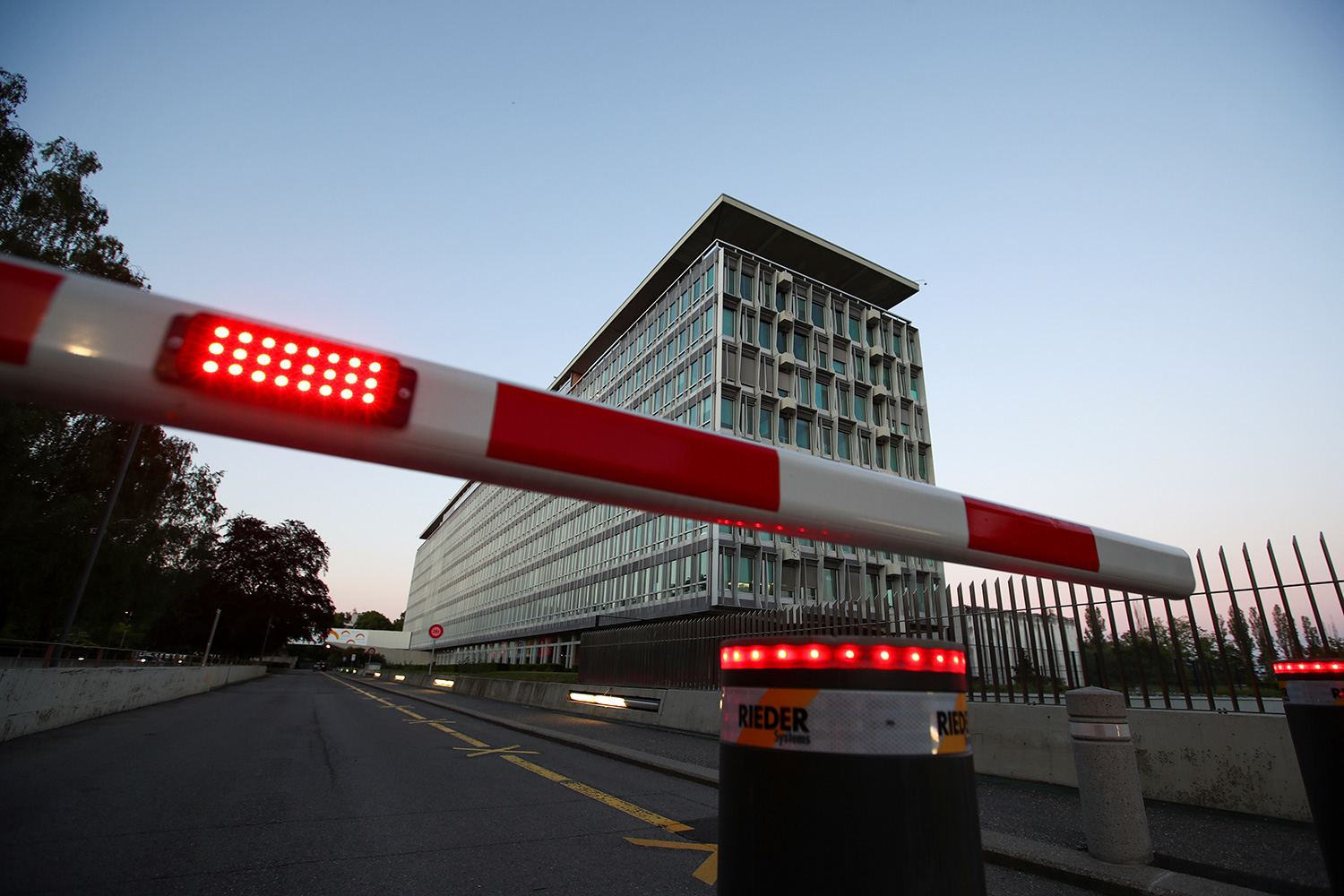 The photo shows the WHO building at night with a red and white guard barrier lowered to block the street entrance.
