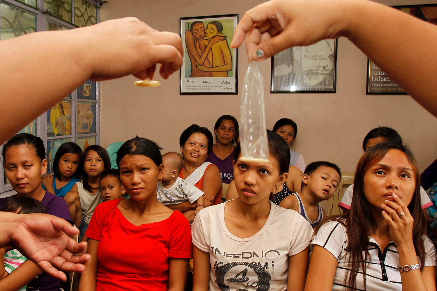 Picture shows the hands of a health worker holding up two condoms—one rolled, one unrolled—in front of a large audience of yound Philipino women.