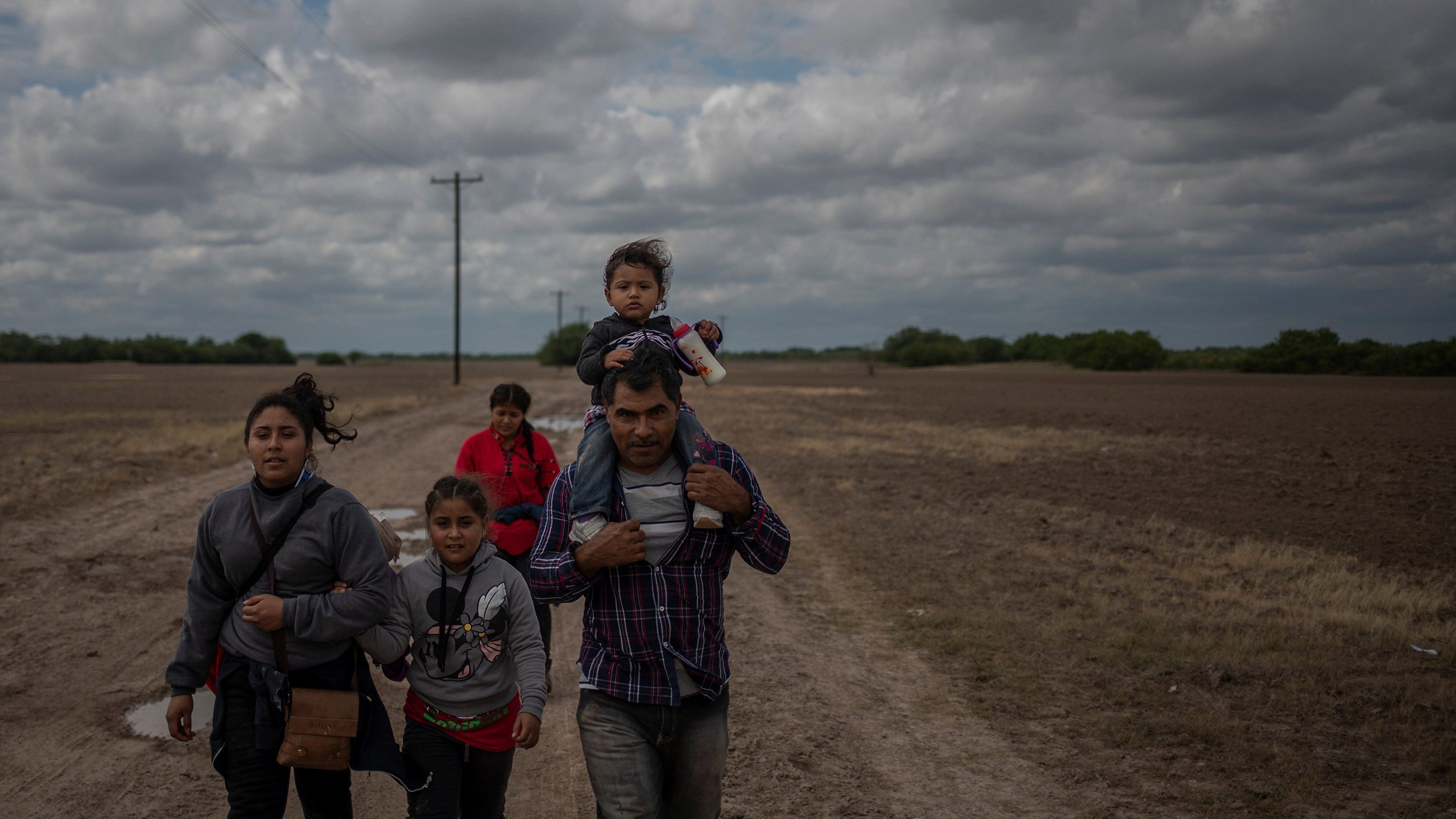 The photo shows a family walking through a field at dusk. The father has a small child on his shoulders.
