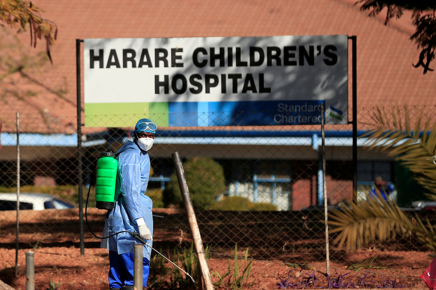 The photo shows a worker in protective gear spraying disinfectant mist in front of a building with a large sign reading, "Harare Children's Hospital.