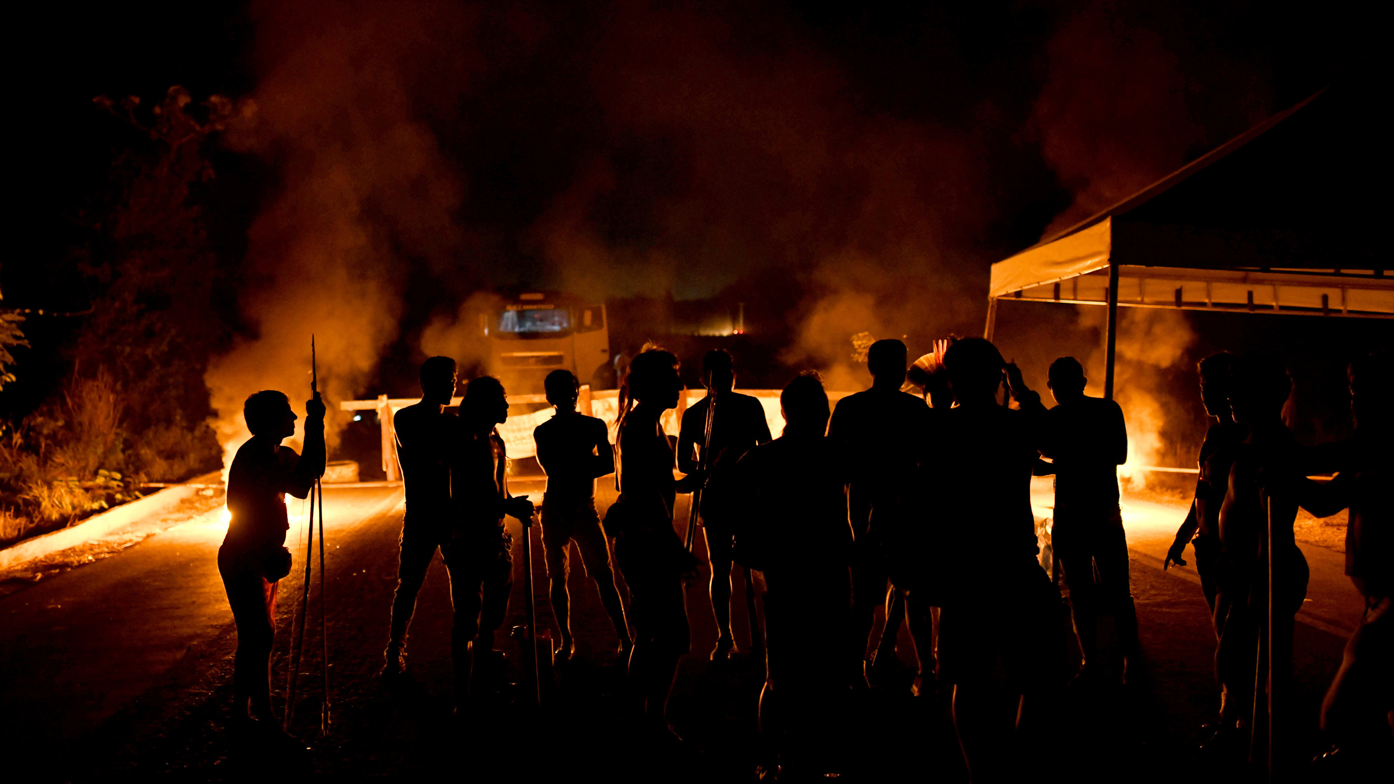 The image shows a large number of protesters at night silhouetted against a fire burning.