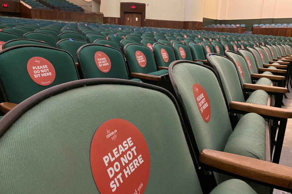 Picture shows row upon row of dull green uncomfortable stadium seating chairs in a nondescript school auditorium. Almost all the chairs are tagged with a sticker that reads, "Please do not sit here."