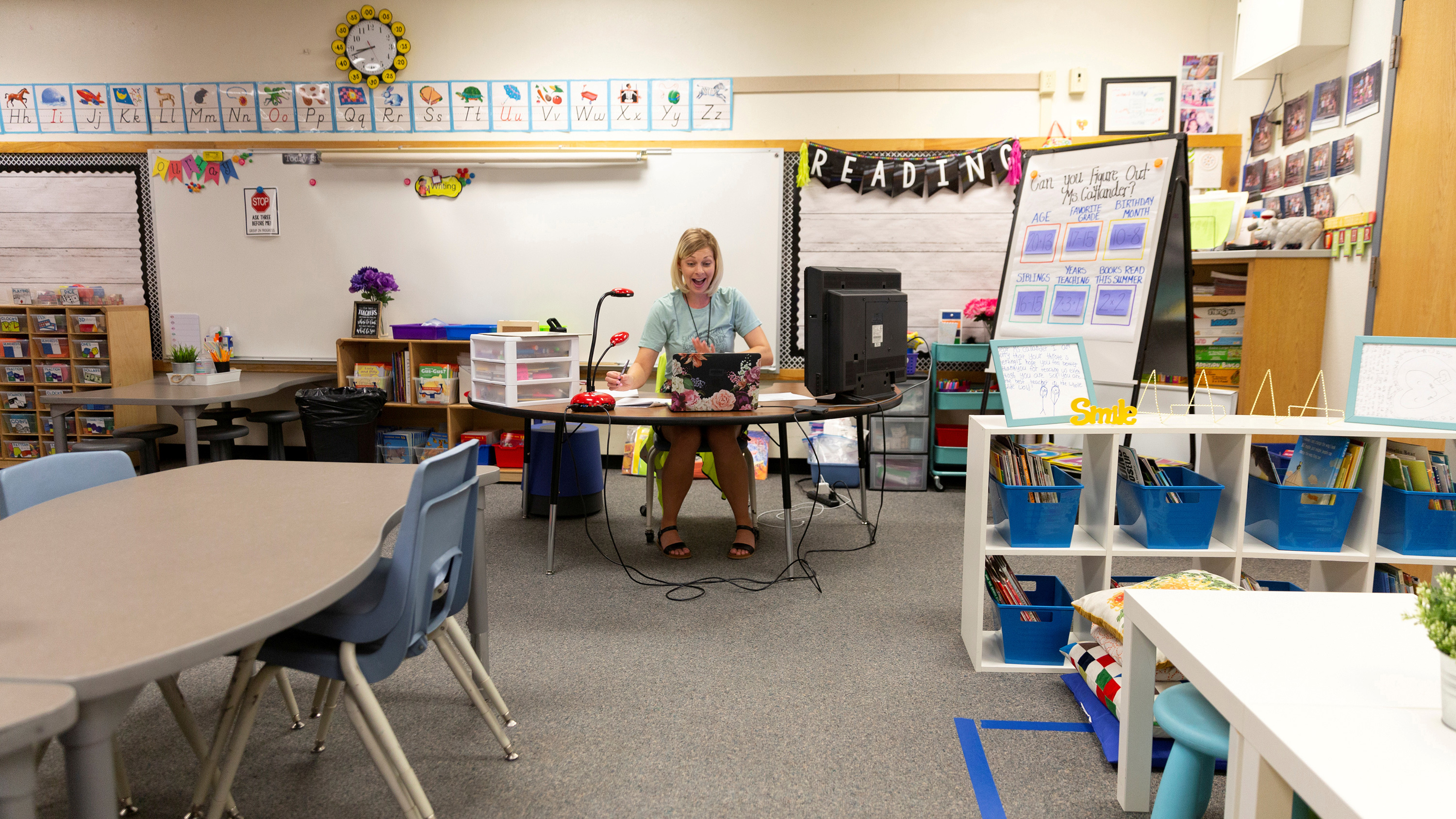 The photo shows the teacher in an empty classroom speaking expressively into the computer.