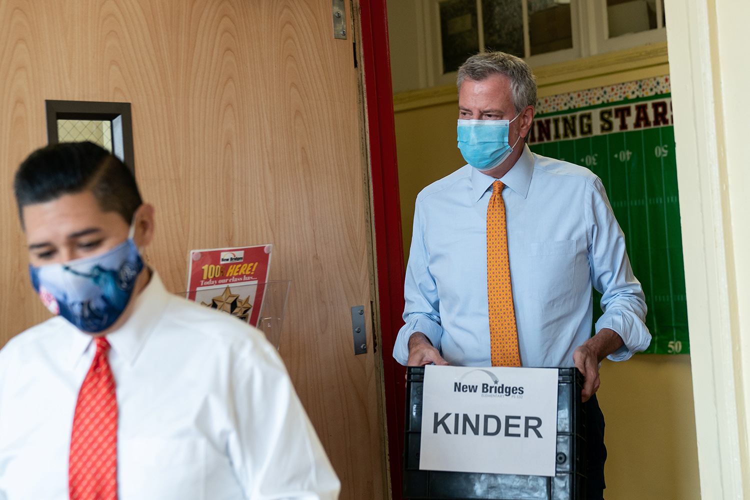 The photo shows the mayor walking into the classroom wearing a mask and carrying a box labeled, "KINDER."