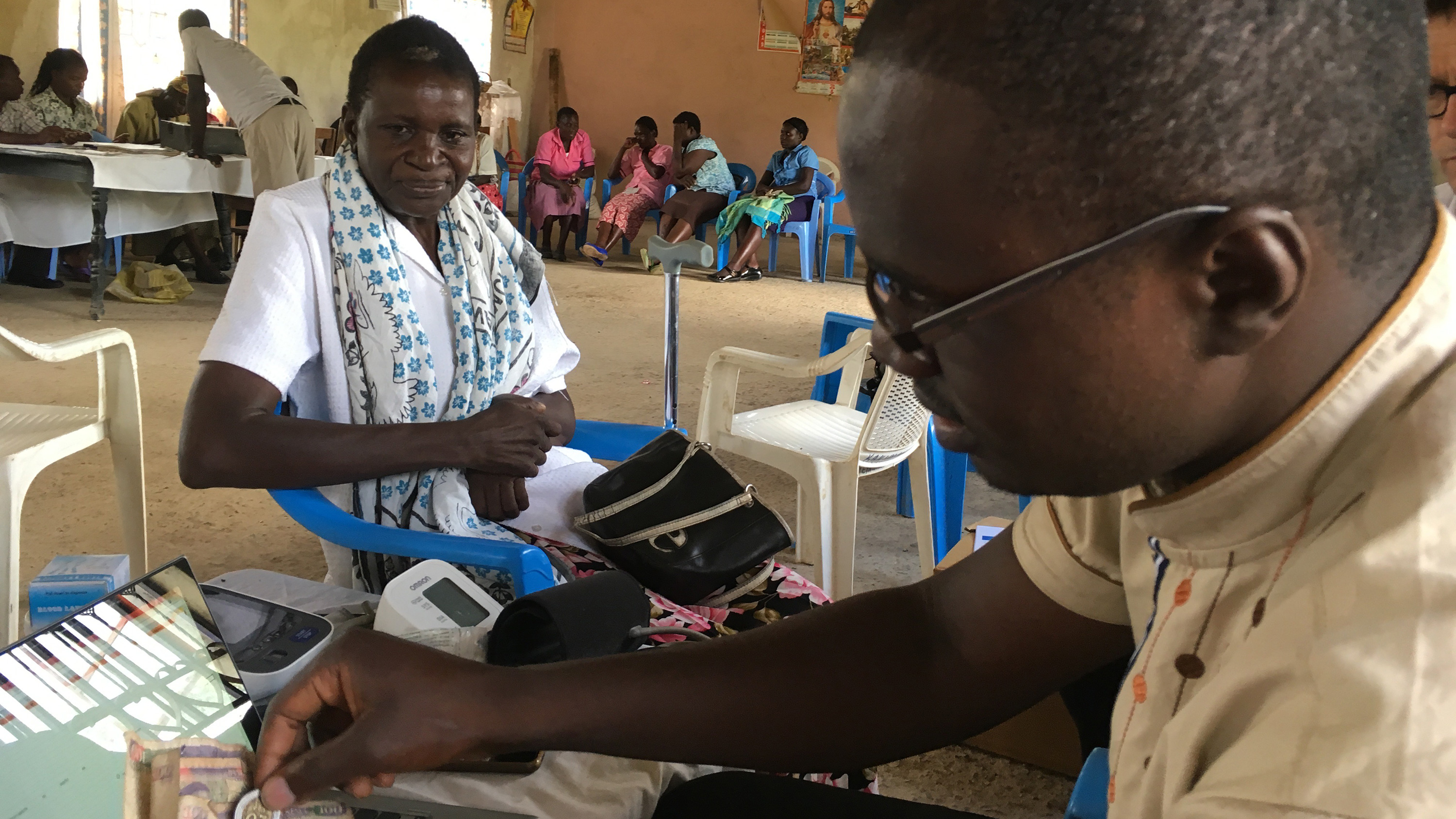 The image shows the interior of a clinic with people waiting in the background while a health worker sits with a patient in the foreground.