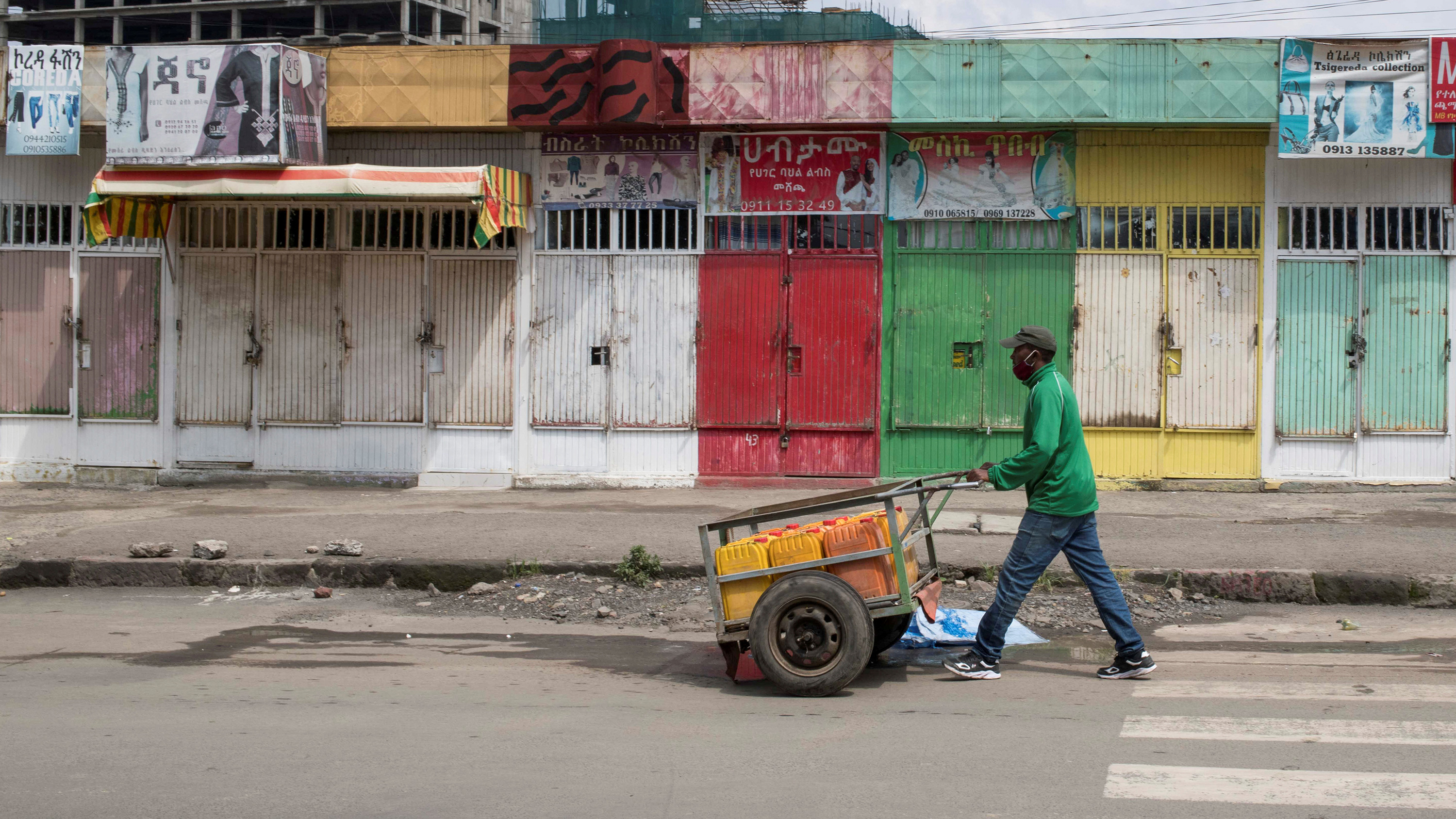 This is a striking photo of a man in a green shirt pushing a cart past wooden shuttered shops painted green and beige and red and yellow.