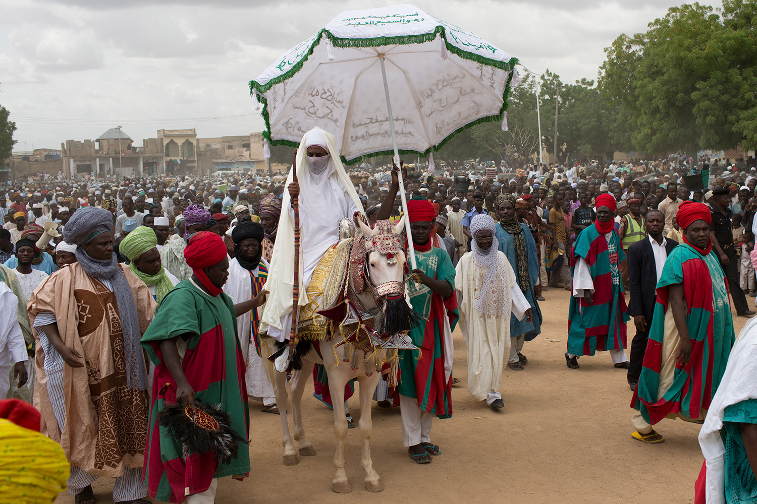 The photo shows the emir atop a horse with decorative coverings while a massive crowd surrounds him and one attendant holds an umbrella high above the emir's head.