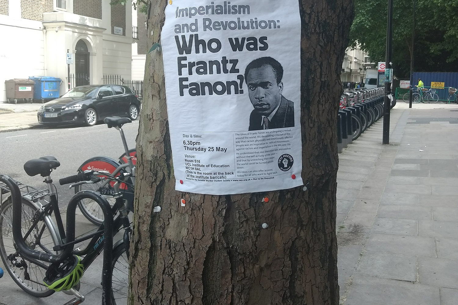 The photo shows a paper flier pasted to a tree that reads, "Who was Frantz Fanon?"