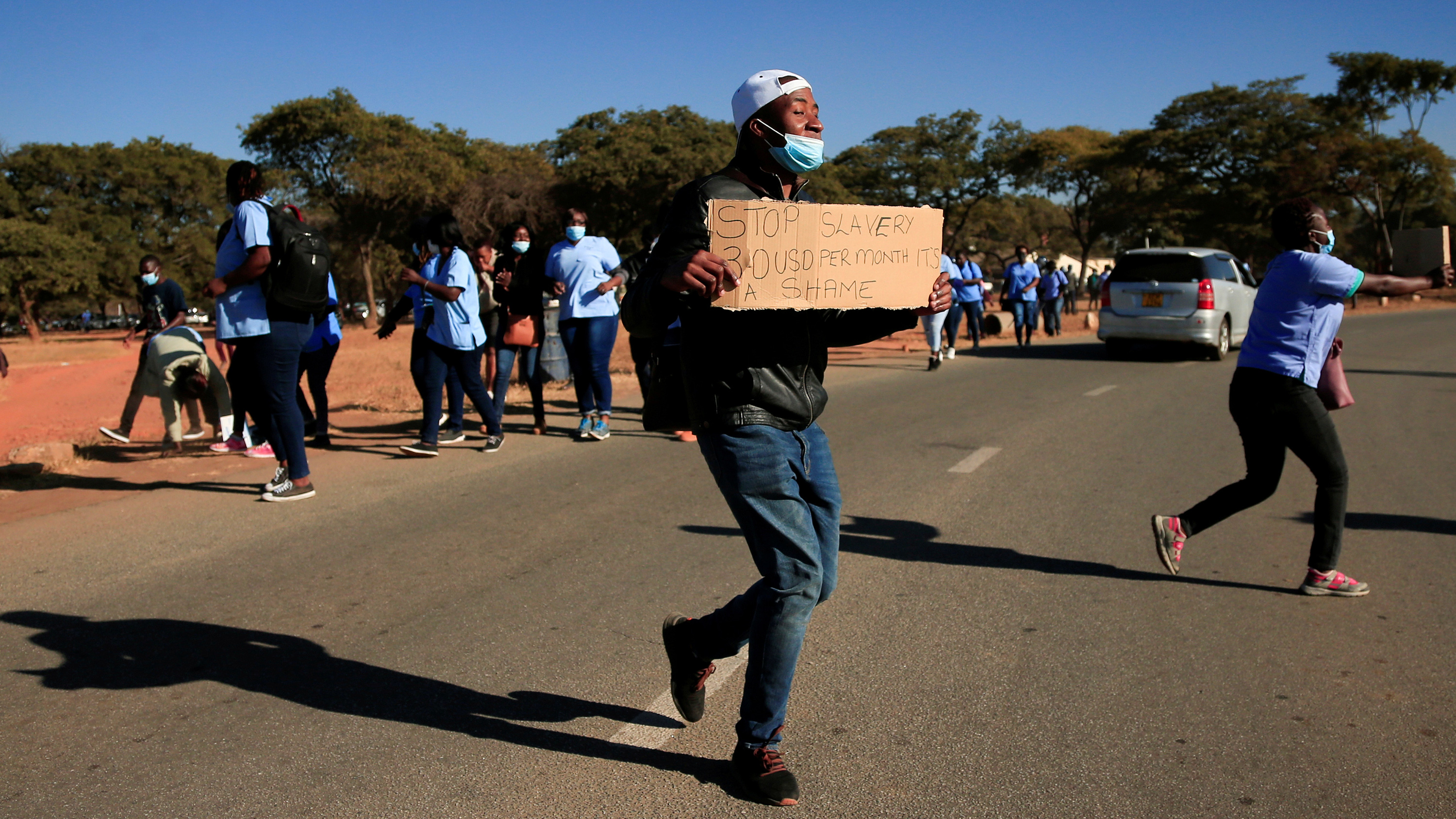 The photo shows a protest of people, one of whom is in the foreground with a hand-painted sign that reads, "Stop Slavery / 30 USD per month / It's a shame."