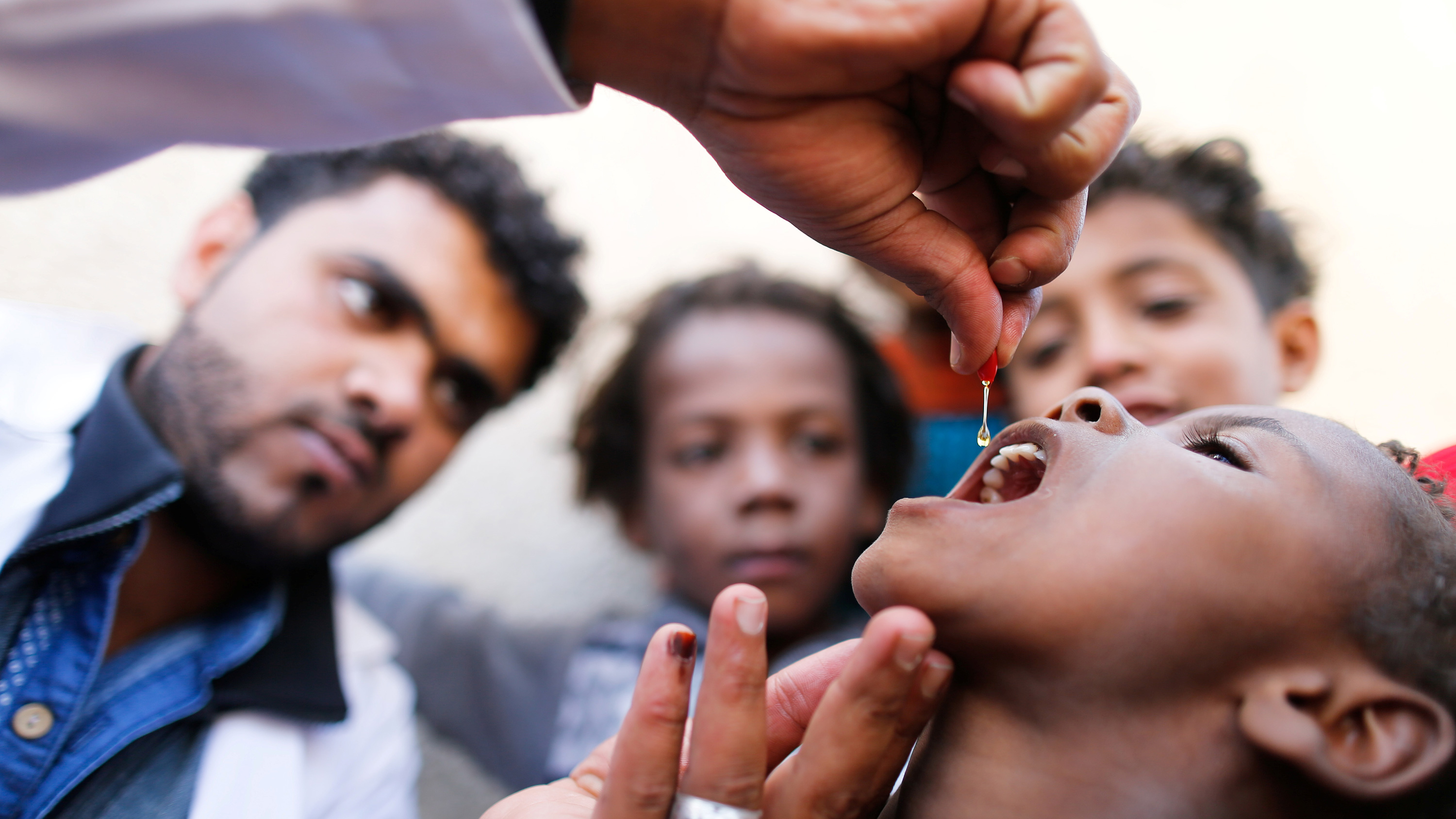 The image shows a health worker holding a dropper above a child's head. He is concentrating on the dropper, from which he is squeezing a clear liquid into the child's open mouth with family members looking on in the background.