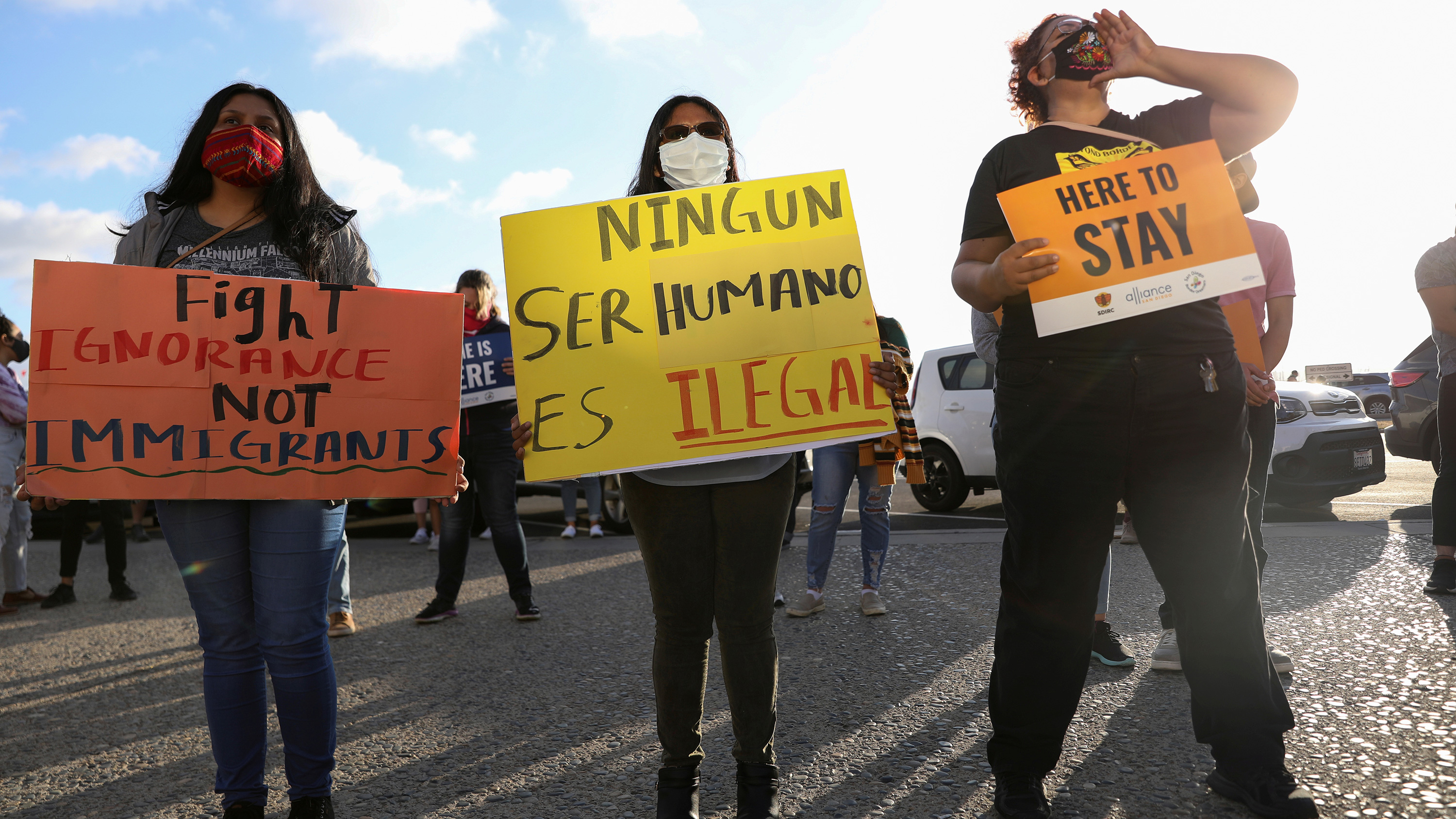 The photo shows three people holding handmade signs in a protest. The sign in the middle reads: "No human being is illegal."