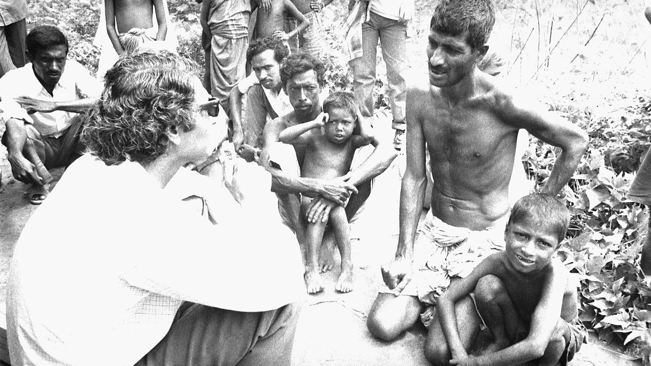 The photo shows the doctor seated on the ground speaking with a number of adults with several children also seated amongst them.