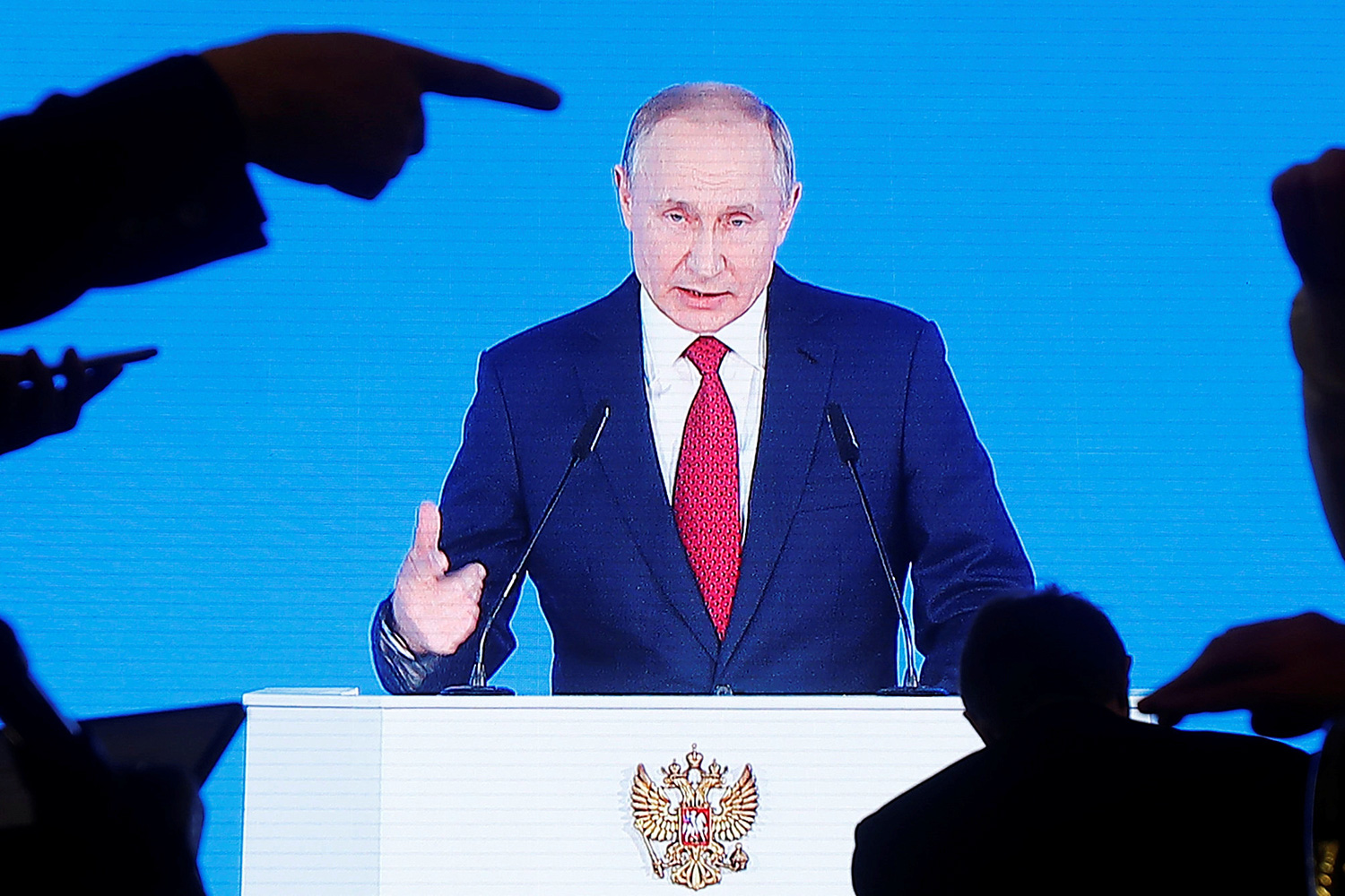 The photo shows the Russian president at a podium speaking into a mic with hands raised in silhouette in the foreground. One appears to be pointing at him, though this is likely an artifact of the camera angle.
